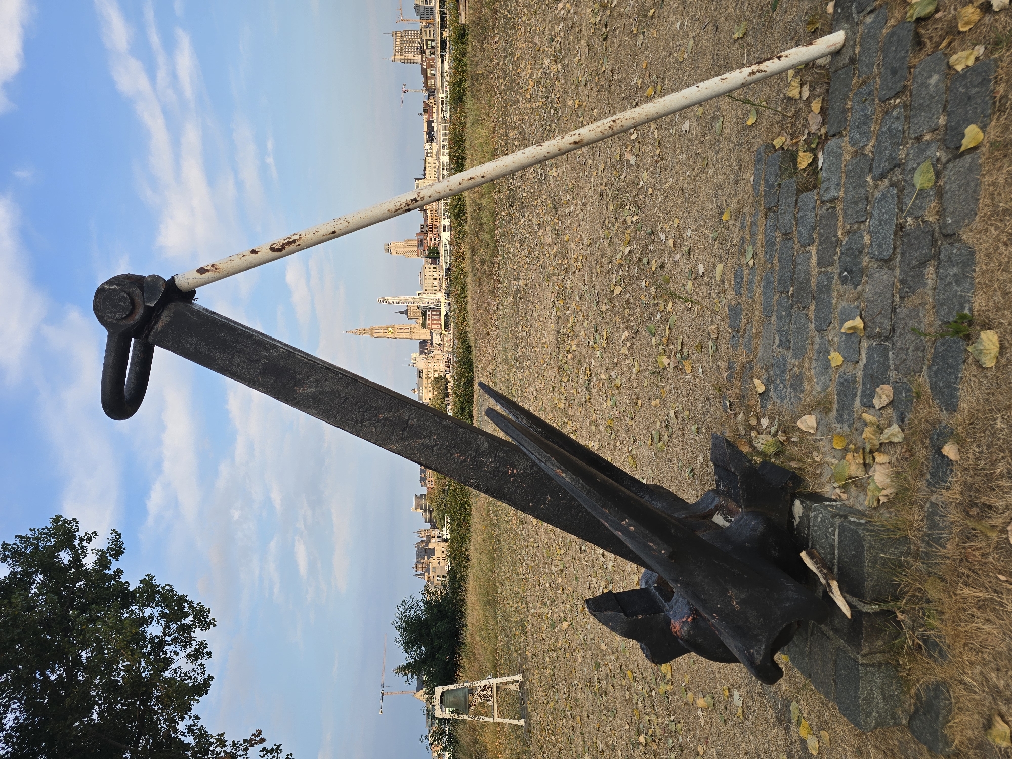 An imposing, weathered black anchor, a relic of maritime history, stands prominently in the foreground of this image, leaning against a rusted white pole. It rests on a base of dark, rough-hewn stones, surrounded by dry, yellowing grass and scattered autumn leaves. In the middle ground, a stark white, A-frame structure supports a bronze bell, adding another layer of historical significance to the scene.
Beyond this, a lush green field stretches towards a distant cityscape. The skyline of Antwerp, Belgium, is clearly visible, dominated by the majestic spire of the Cathedral of Our Lady, a Gothic masterpiece, and a Ferris wheel, a modern contrast to the ancient architecture. The city's waterfront is lined with buildings of various architectural styles and heights, and a river flows in front of it. Cranes, industrial structures indicative of a busy port, dot the horizon. The sky above is a vibrant blue, adorned with scattered white clouds, suggesting a pleasant, possibly late afternoon, atmosphere. The overall impression is one of a historical monument set against a backdrop of a living, breathing city, where past and present coexist.