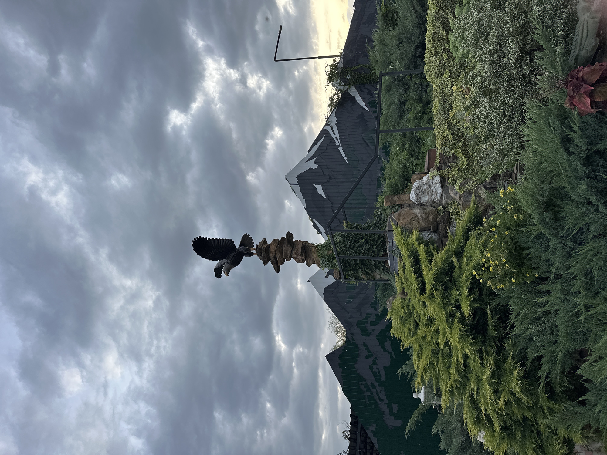 The image depicts an outdoor scene, likely a park or decorative garden area, with a sculpture of an eagle perched atop a stack of stones. In the background, there are stylized mountain-like structures made of painted wood or metal, suggesting a themed environment. Lush green vegetation, including bushes and trees, fills the foreground and middle ground. The sky is overcast with dramatic, cloudy formations, hinting at either pre-storm conditions or twilight. The overall impression is of a man-made landscape designed for aesthetic appeal, possibly within a public space. The location is identified as Yaroslavskaya, Russia.