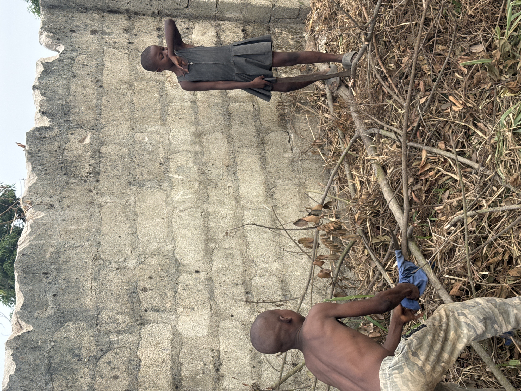 The image captures two young individuals in an outdoor setting within Ihiagwa, Nigeria, against a backdrop of a weathered concrete block wall with an uneven top edge. The ground is covered with a dense layer of dry branches, fallen leaves, and sparse green vegetation.
In the left foreground, a shirtless individual, appearing male, is positioned from the back, bending forward. This individual wears camouflage-patterned trousers and is engaged in handling a cluster of dry branches, holding a blue fabric item in their hands.
To the right, a second individual, appearing female, stands upright, wearing a dark grey sleeveless dress with a pleated skirt and light-colored sandals. Her right hand is raised, touching the area near her nose and mouth. She holds a long stick vertically in her left hand, with the lower end resting on the ground.