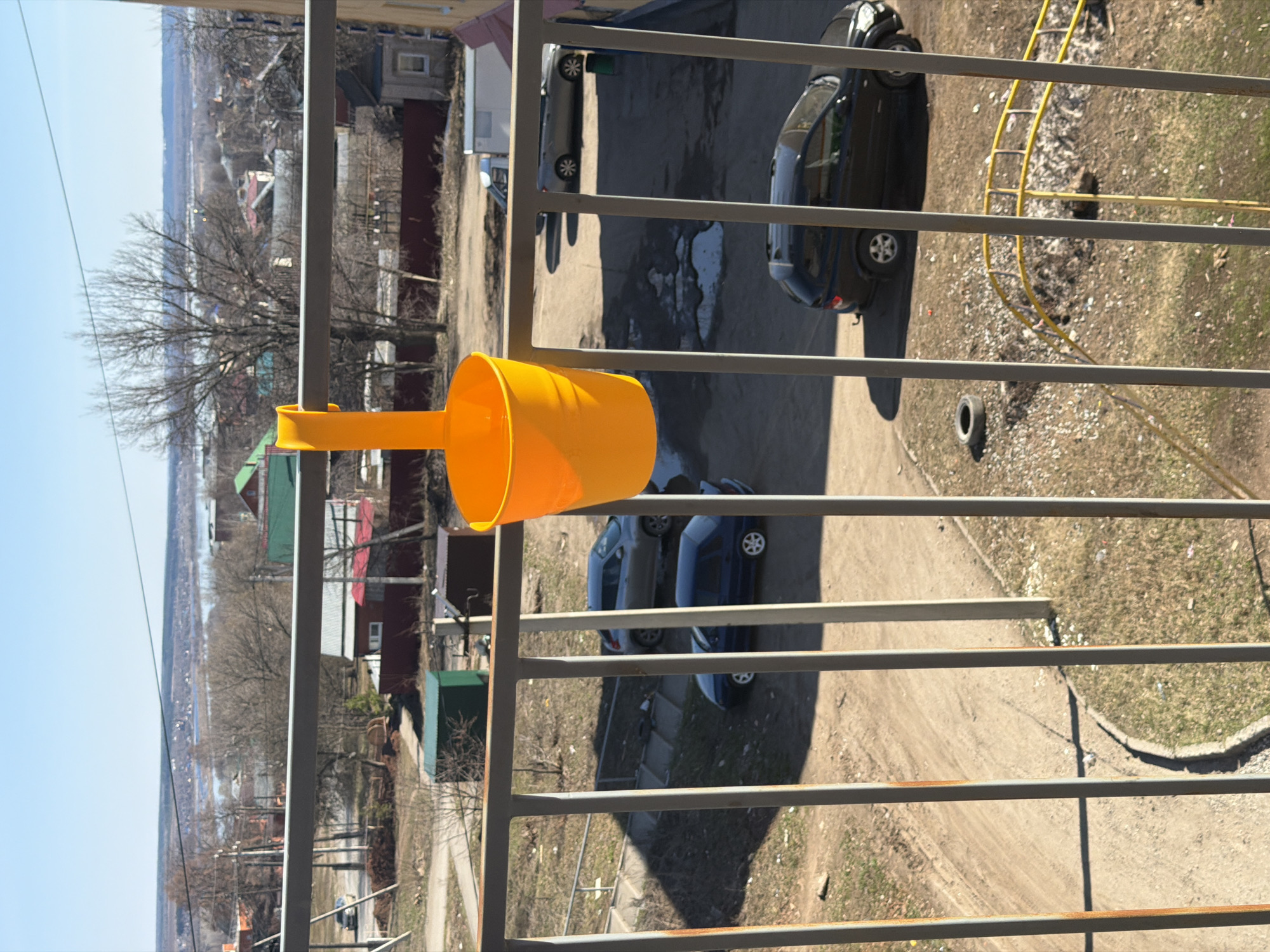 The image captures an outdoor scene, likely viewed from an elevated position such as a balcony, looking down and across a residential area. In the immediate foreground, a bright yellow plastic funnel or scoop is attached to a gray metal railing, which shows signs of rust in places.
Beyond the railing, the view extends over an unpaved, dirt ground that serves as a parking area. Several cars are visible: a dark-colored sedan is parked on the right, and further left, a silver hatchback and a blue sedan are also parked. The ground is dry, with patches of sparse grass and scattered debris, including a discarded tire and a yellow flexible pipe or hose near the dark sedan. The presence of strong, elongated shadows suggests a sunny day, likely mid-morning or afternoon, under a clear blue sky.
In the mid-distance, numerous trees with bare branches indicate that the season is late winter or early spring. Scattered among these trees are various residential buildings with different colored roofs, including red and green. Further in the background, a wide, lighter-colored expanse is visible, possibly a river or a large body of water, which could be partially frozen or reflecting the sky. Distant hills or land formations frame the horizon. A faint wisp of smoke or haze can be seen above the distant trees. The overall setting depicts a somewhat semi-urban or rural residential landscape, consistent with Sarapul, Russia. There are no people visible, and no discernible text. The scene is static, offering a serene observation of the surroundings.