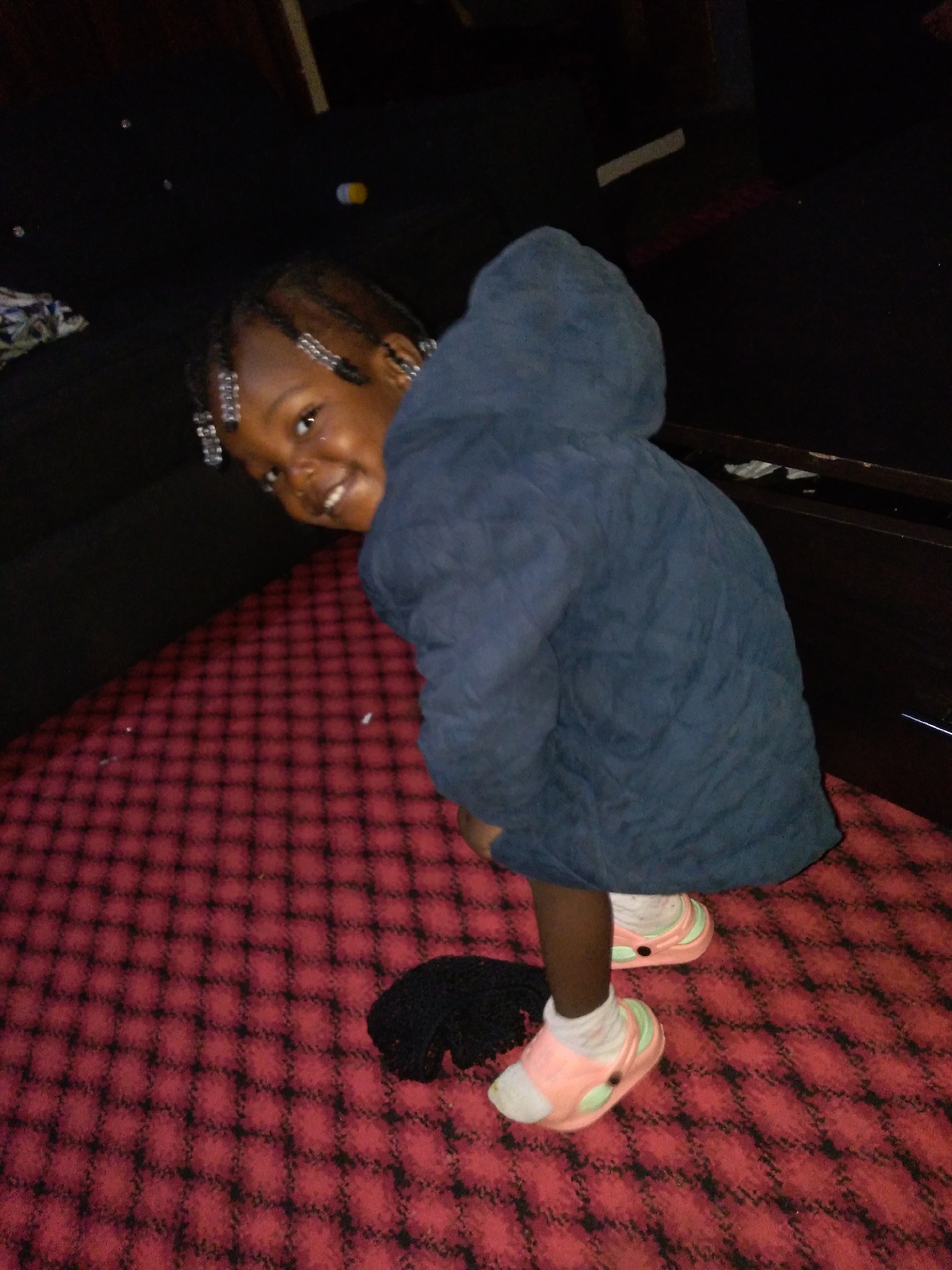 A young Black girl with braided hair adorned with silver beads is captured in a medium shot, bending down and looking towards the camera with a wide smile. She is wearing a dark blue quilted puffer jacket and pink and mint green Crocs with white socks. The girl is positioned on a red carpet with a black checkered pattern. To the right of the girl, a dark wooden dresser or cabinet is visible. The background is dimly lit and appears to be indoors. The scene is set in an indoor environment, possibly a bedroom or living area, in an unspecified location.