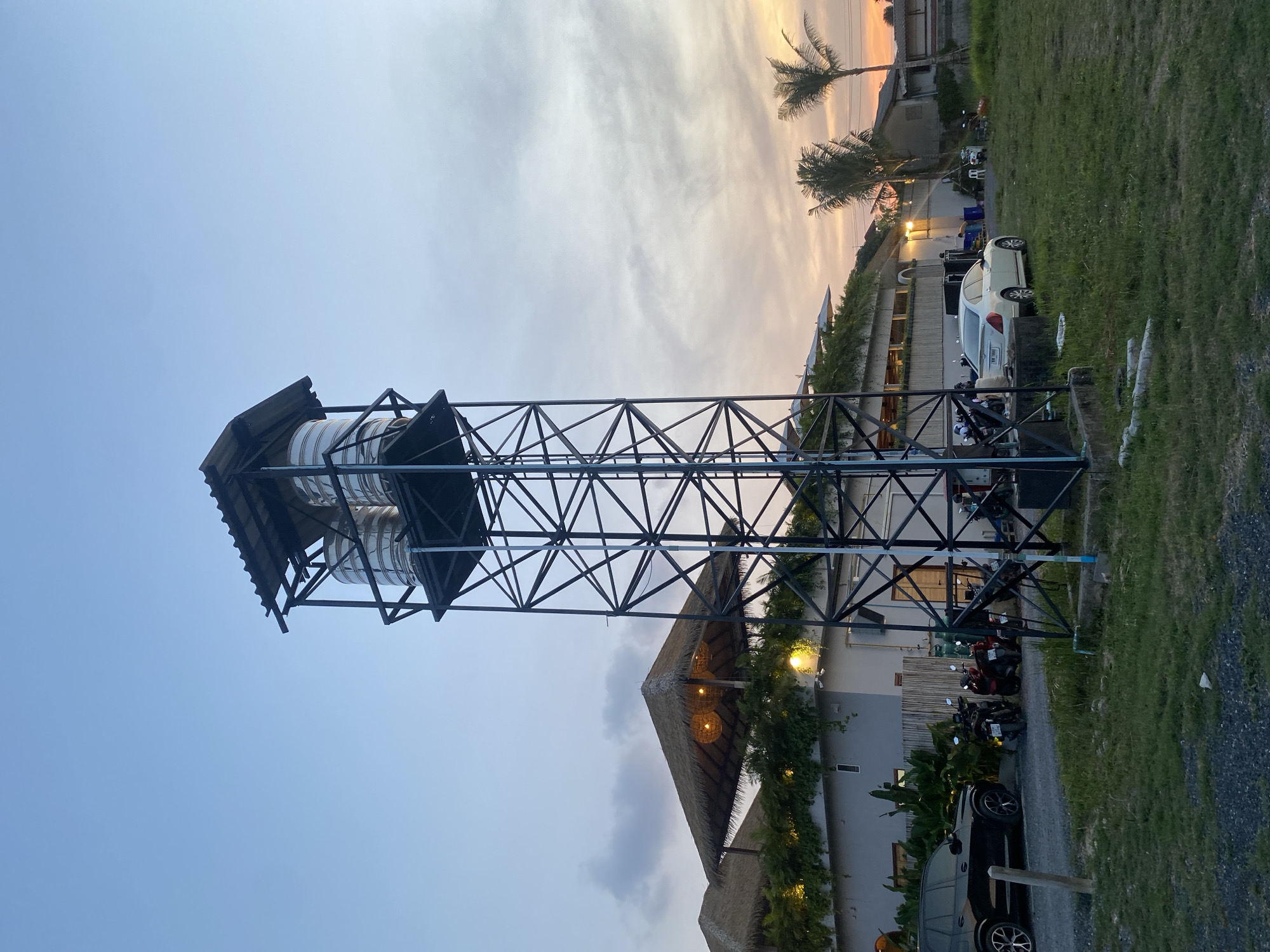 The image is taken in Choeng Thale, Thailand, and features a metal water tower with multiple cylindrical tanks near a building with a thatched roof. The tower is constructed of black metal beams in a latticework pattern. Palm trees are visible in the background, silhouetted against the sky with hues of orange and pink at dusk. In front of the building, there are several motorcycles and at least two cars parked, including a white sedan and a black SUV. A grassy area is located on the right side of the frame. The lighting suggests it's late afternoon or early evening.