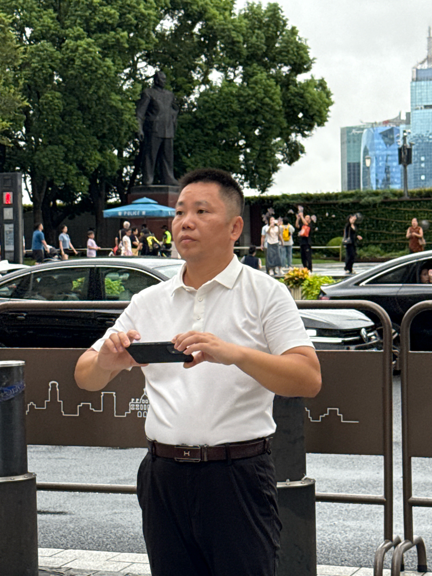 A middle-aged East Asian man wearing a white polo shirt and black pants stands in the foreground, holding a black smartphone in both hands, appearing to take a picture. Behind him, a blurred urban scene unfolds. A large, dark-colored car is parked to his left, and another dark-colored car is visible further back. In the mid-ground, several people are walking and standing near a monument featuring a statue of a man, with a blue umbrella labeled "POLICE" behind it. Trees with lush green foliage are scattered throughout the scene. In the background, modern skyscrapers with reflective glass facades rise against a cloudy sky. The overall setting suggests a public space in Shanghai, China, possibly a park or plaza with significant historical and architectural elements.