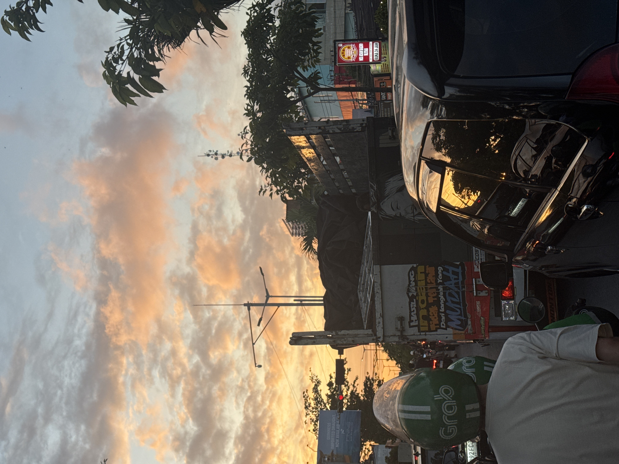 The image depicts a traffic jam on a street in a city at sunset. The sky is filled with orange and pink clouds, reflecting the warm glow of the setting sun. In the foreground, a person wearing a green Grab helmet is visible from the back, seated on a motorcycle. To their right, a black car's side window reflects the sunset. In the middle ground, a truck is stopped in traffic, with various stickers and graffiti on its side. The truck is laden with a dark tarp. Beyond the truck, more vehicles are visible, suggesting a heavy flow of traffic. Traffic lights, streetlights, and utility poles are also present, indicating an urban environment. Buildings and trees are seen in the background, partially obscured by the traffic and the sunset's haze. The overall impression is one of a typical evening commute in a bustling city, with the dramatic sunset adding a picturesque element to the scene.