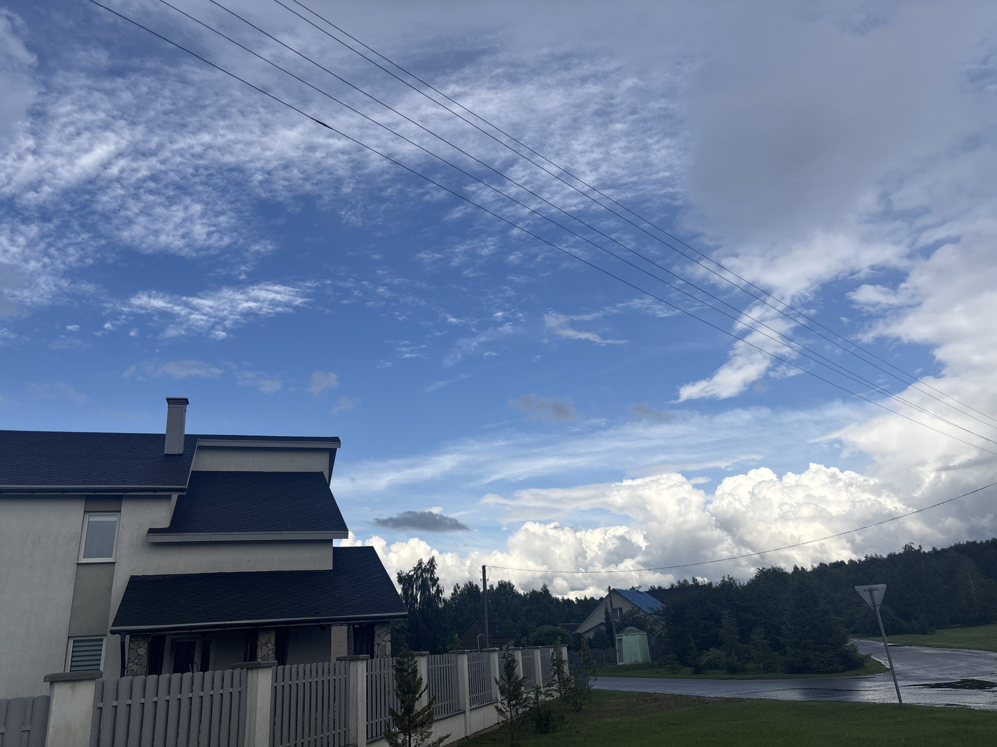 The image depicts a residential scene under a dynamic sky, likely in the Капыльскі сельскі Савет, Belarus. In the foreground, a portion of a modern house with a dark, multi-pitched roof and light-colored walls is visible on the left. A grey, picket-style fence runs across the front of the property. Behind the house, a dense line of dark green trees forms the background, obscuring the horizon in many areas. Beyond the trees, several residential structures are sparsely visible, including one with a blue roof and a smaller, light green shed-like building next to it. The sky is a prominent feature, with a bright blue expanse partially filled with scattered, wispy white clouds and larger, billowy cumulus clouds on the right. Four dark power lines stretch diagonally across the upper portion of the frame, from left to right. The road in the mid-ground appears wet, suggesting recent rainfall, with puddles visible. A triangular road sign is planted on the right side of the road. The overall atmosphere suggests a rural or suburban setting with evidence of overcast and potentially clearing weather.