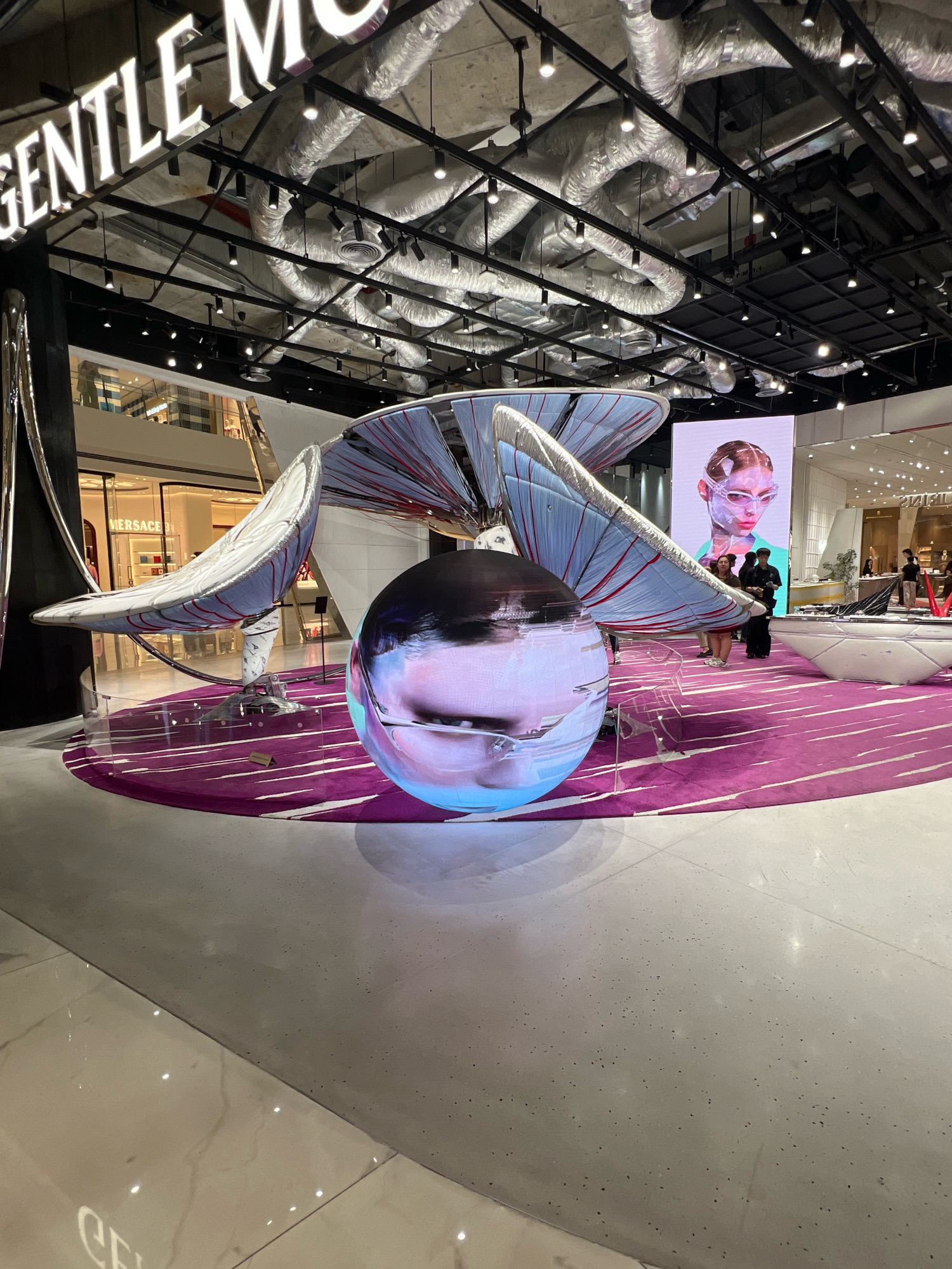 This image captures a display within a retail environment, possibly a shopping mall, featuring a large, abstract sculpture. The sculpture, with its fan-like elements in blue and red, is illuminated and stands on a purple rug. A spherical display screen in the foreground shows a close-up of a person's face, distorted by the curvature of the sphere. Behind the sculpture, a large digital screen displays a portrait of a person wearing futuristic eyewear, their face also partially obscured by a transparent layer, suggesting a theme of technology or augmented reality.
Several people are visible in the background, observing the display. One individual in a black hat and dark clothing appears to be an attendant or security personnel. The setting includes elements of a modern commercial space with exposed ductwork and lighting visible on the ceiling. The floor is a mix of polished marble and a grey, textured material. Storefronts are visible in the background, one of which has "VERSACE" written on it, indicating a luxury retail area. The overall impression is one of a contemporary and visually striking promotional display, likely for fashion or technology. The lighting suggests it is daytime, and there are no indications of weather conditions. The location is identified as Bangkok, Thailand.