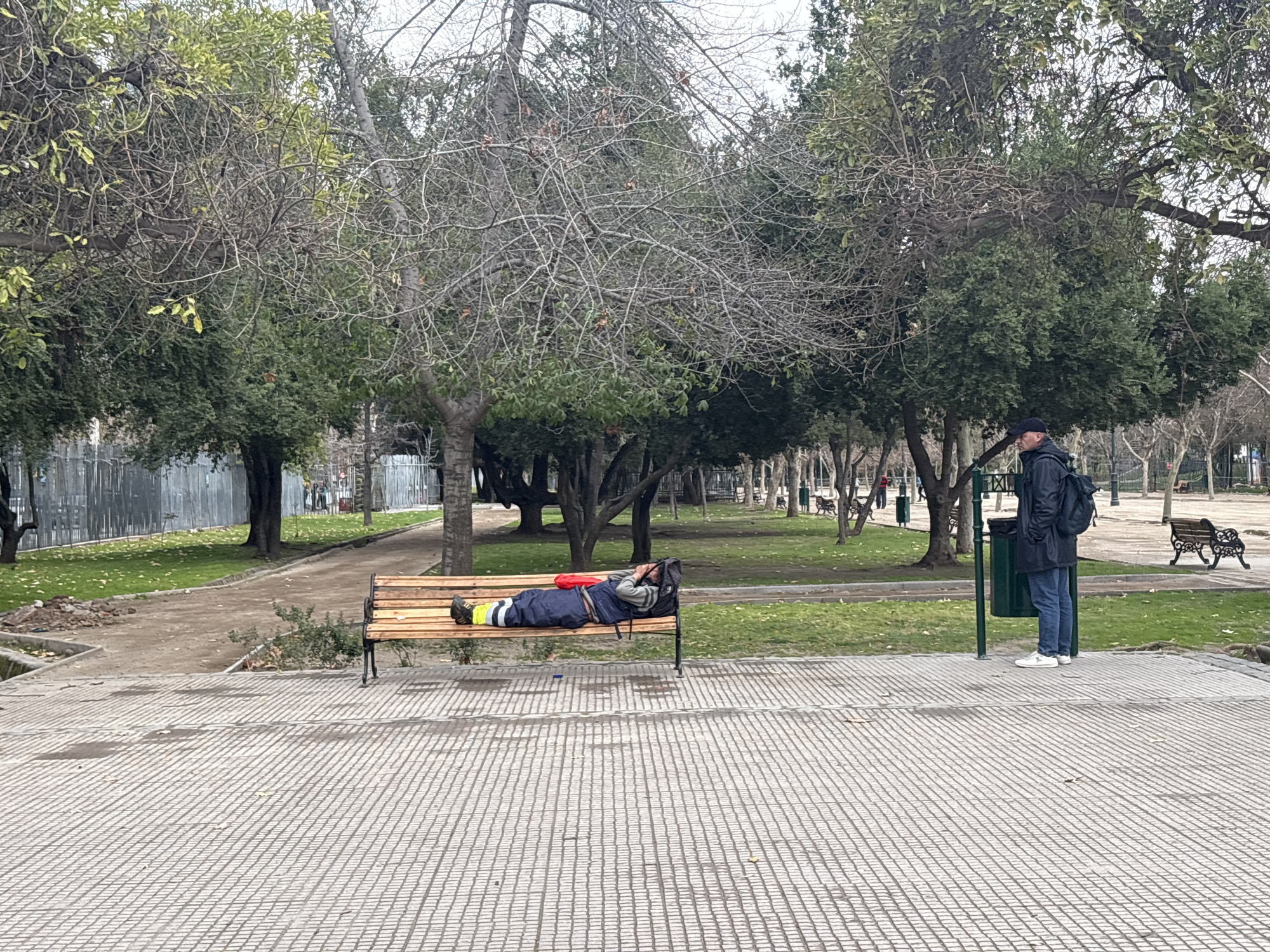 A man wearing a black jacket and backpack stands near a green trash receptacle in Parque Forestal, Santiago, Chile. A second person, dressed in a blue jumpsuit and a yellow vest, sleeps on a wooden park bench. The park features mature trees with sparse foliage, a paved pathway, and a grassy area.