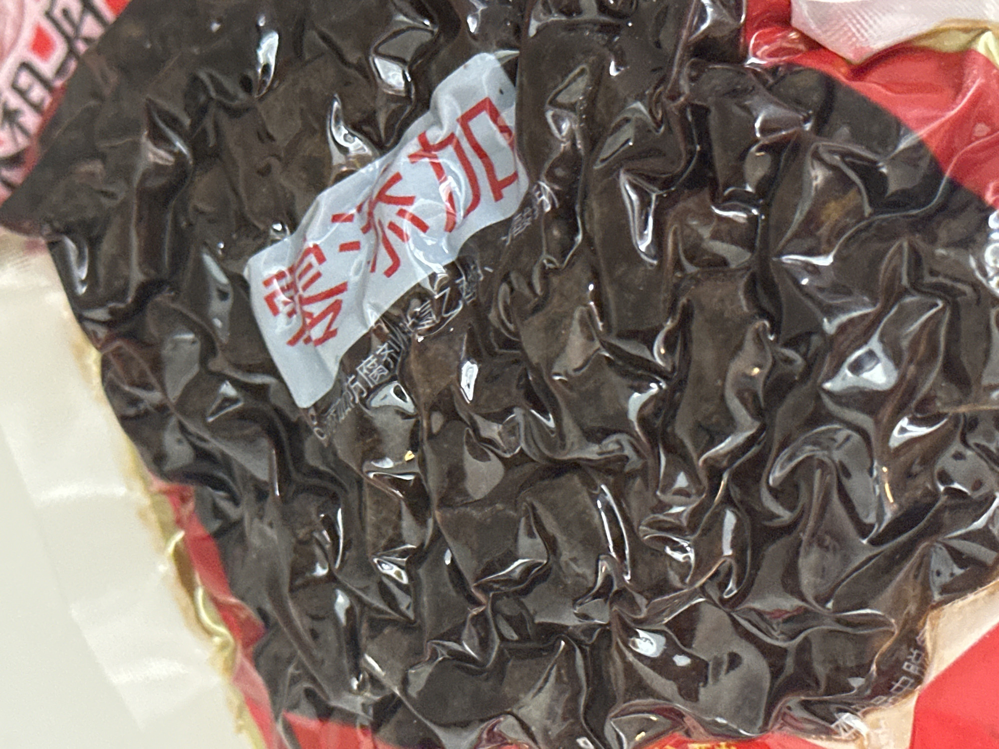 The image shows a close-up, overhead view of a vacuum-sealed package of dark brown, possibly food-related, items. The packaging is crinkled and reflects light, obscuring some of the details of the contents. A small white label with red Chinese characters is affixed to the top of the packaging. The background is out of focus, with hints of red and white colors. There is no visible information indicating the location in Nevinnomyssk, Russia.