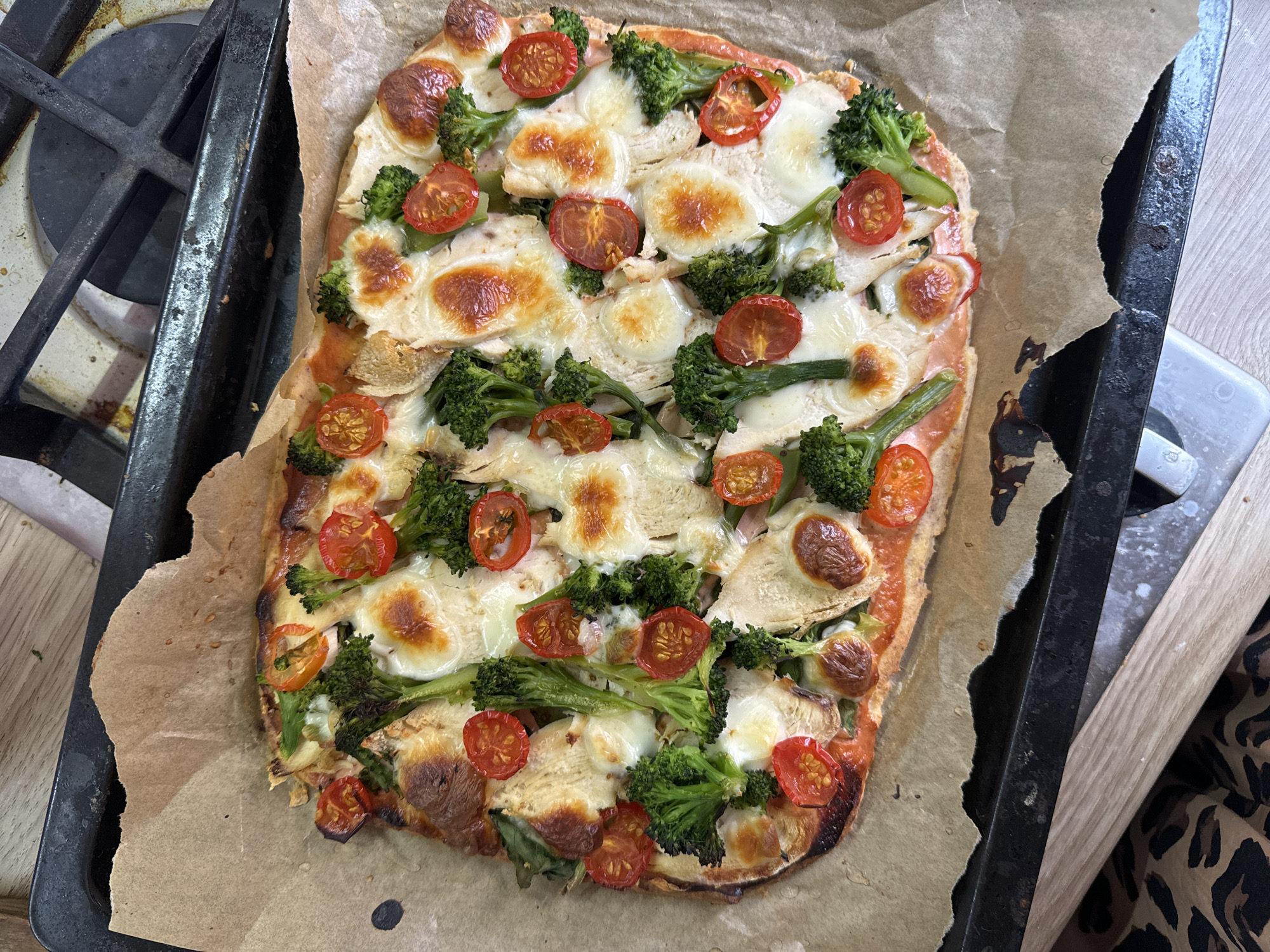 A rectangular pizza on a baking sheet is positioned next to a gas stovetop. The pizza has a browned crust and is topped with tomato sauce, mozzarella cheese, broccoli florets, and sliced cherry tomatoes. The baking sheet is lined with parchment paper and sits on a black metal surface. The gas stove is black with metal grates. A patterned fabric is visible on the right side of the image.