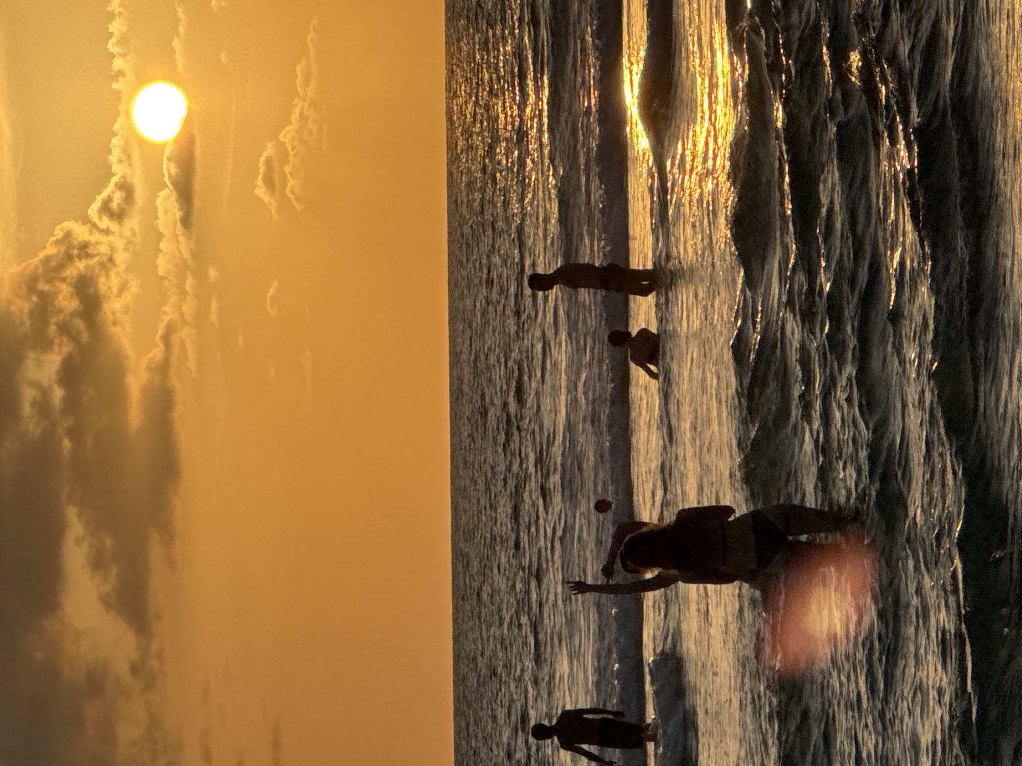 A vertical, low-angle, full-shot photograph captures silhouettes of four people in the ocean at Karon Beach, Thailand, during a sunset. The sun, a bright, white orb, is positioned in the upper right quadrant of the image, partially obscured by thin, wispy clouds. The sky is a gradient of orange and yellow, transitioning to a darker orange near the horizon. The ocean is a deep blue-gray with visible waves, reflecting the sunset's light in shimmering streaks across the surface.
In the foreground, a person with long hair, seen from the back and wearing a bikini, is partially submerged in the water. Their left arm is raised with the index finger pointing upwards. To their left, another person, also from the back, stands with their arms down, facing away from the camera. To the right of the first person, a third person is visible as a silhouette, standing in the water and looking towards the left. In the mid-ground, a fourth person stands in the water on the far left of the frame, facing to the right. All individuals are silhouetted against the bright sky and ocean. A small, dark sphere, possibly a ball, floats in the water between the two central figures. The overall impression is one of leisure and recreation during a tropical sunset.