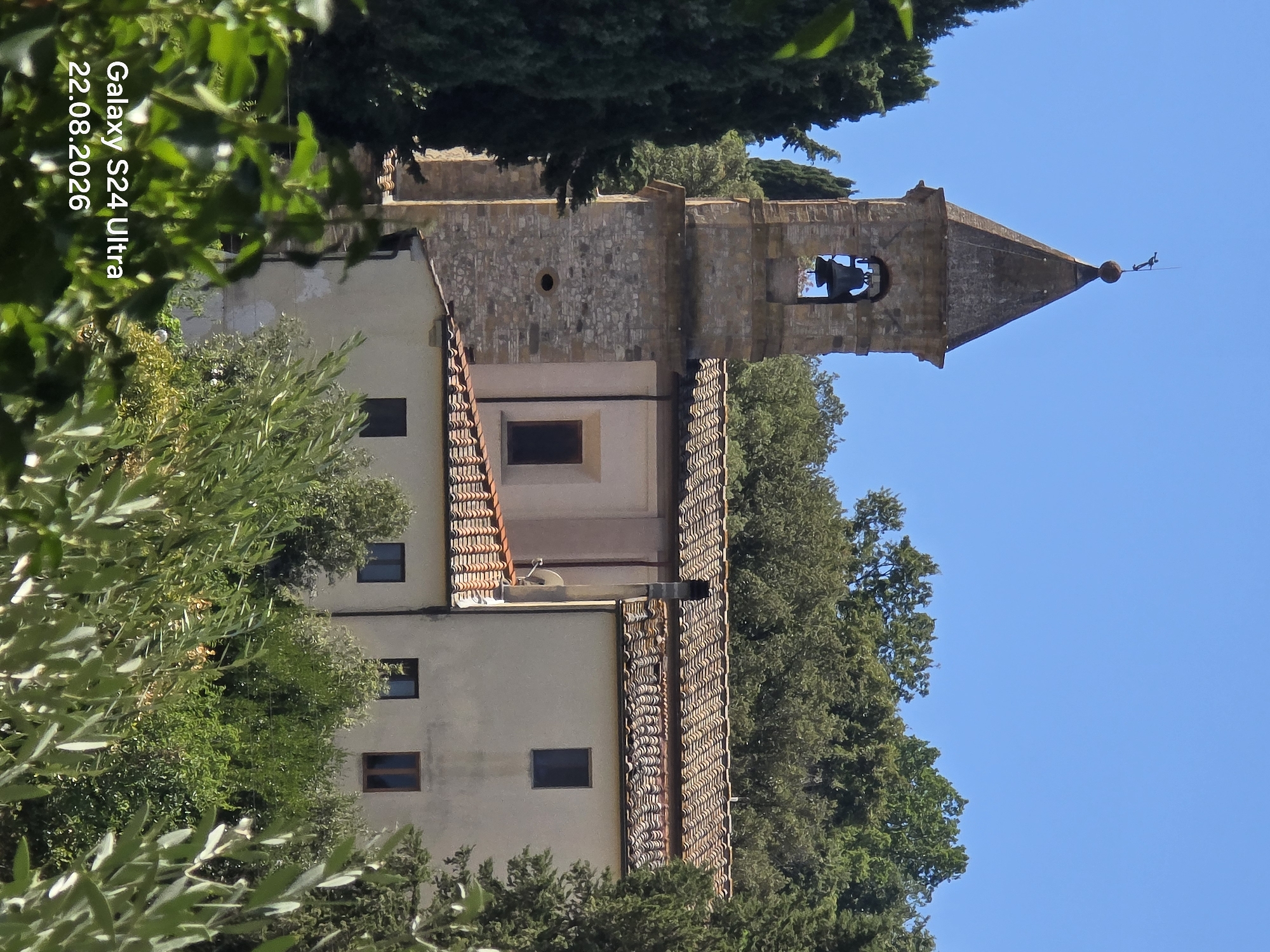 An outdoor, daytime, medium shot features a bell tower of a rustic stone and stucco building, possibly a church, situated in Orciatico, Italy. The tower, with a dark, pyramidal roof topped by a weather vane, has a bell visible within an open archway. Attached to the tower is a cream-colored structure with a red tile roof. The building is partially obscured by lush green trees and foliage in the foreground and surrounding the structure. The sky is a clear, bright blue. A watermark in the bottom left corner reads "Galaxy S24 Ultra 22.08.2026".