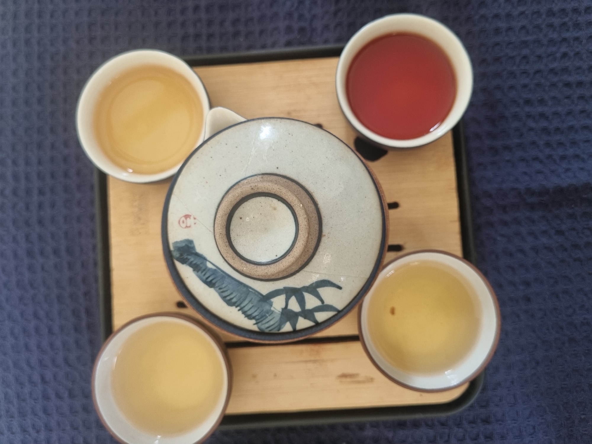 On a dark blue textured surface, a light-colored wooden tray with a black border holds a tea set. The set includes three cups filled with a light-colored beverage and one cup with a red beverage. A circular teapot lid with a grey-blue floral design and a brown center sits in the middle of the tray. The scene appears to be in a domestic setting. There is no indication of it being located in Pedda Kammavaripalli, India. There are no people in the image.