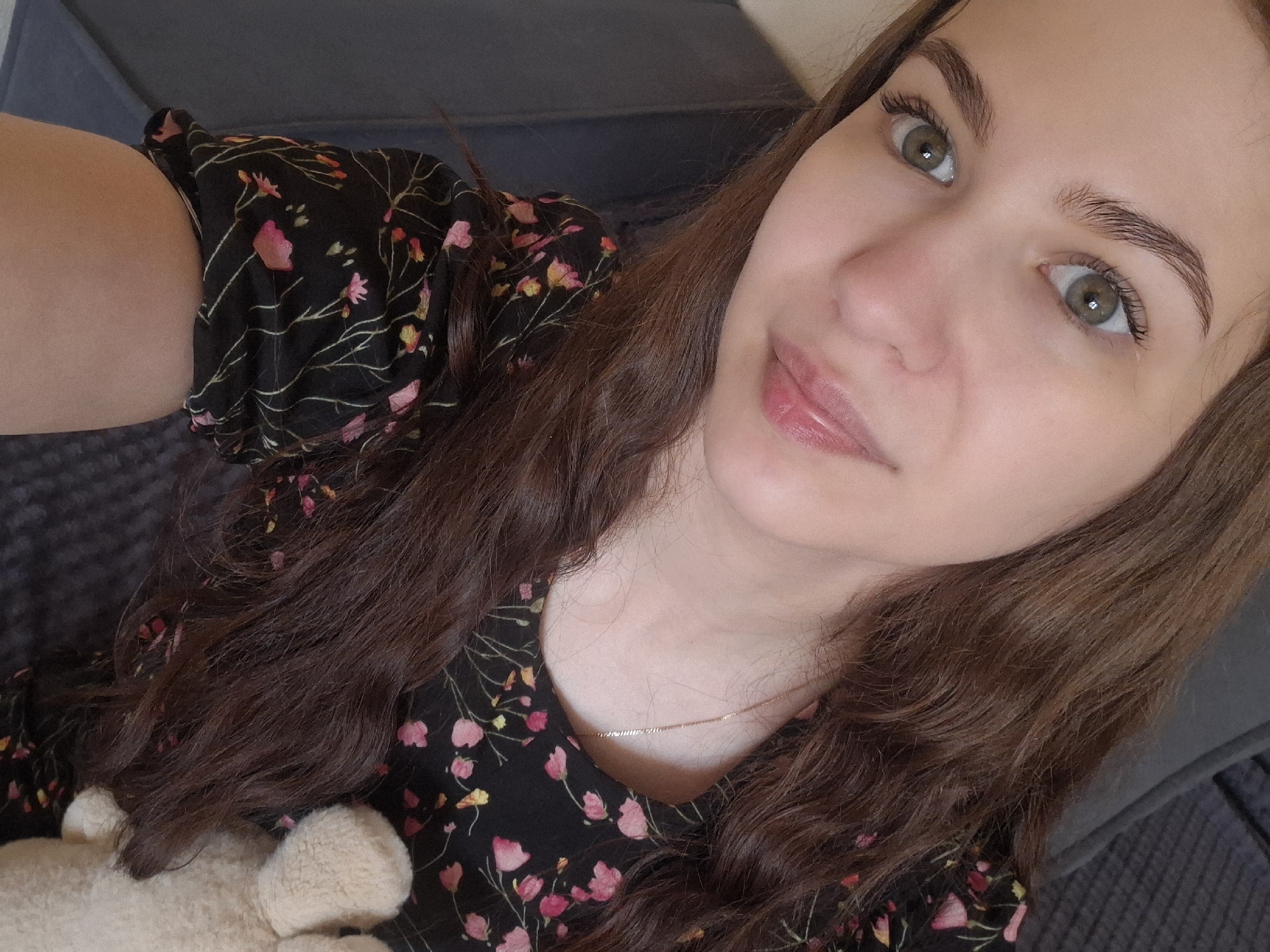 A woman with light skin and green eyes is positioned in a close-up selfie. She has dark brown, wavy hair that falls around her shoulders and is wearing a dark floral-print dress. A portion of a light-colored, soft object, possibly a stuffed animal, is visible near her left shoulder. A thin gold necklace is visible around her neck. The background consists of a gray, textured surface, likely a piece of furniture. There is no indication of the location being in Chaikovsky, Russia, or any specific location.