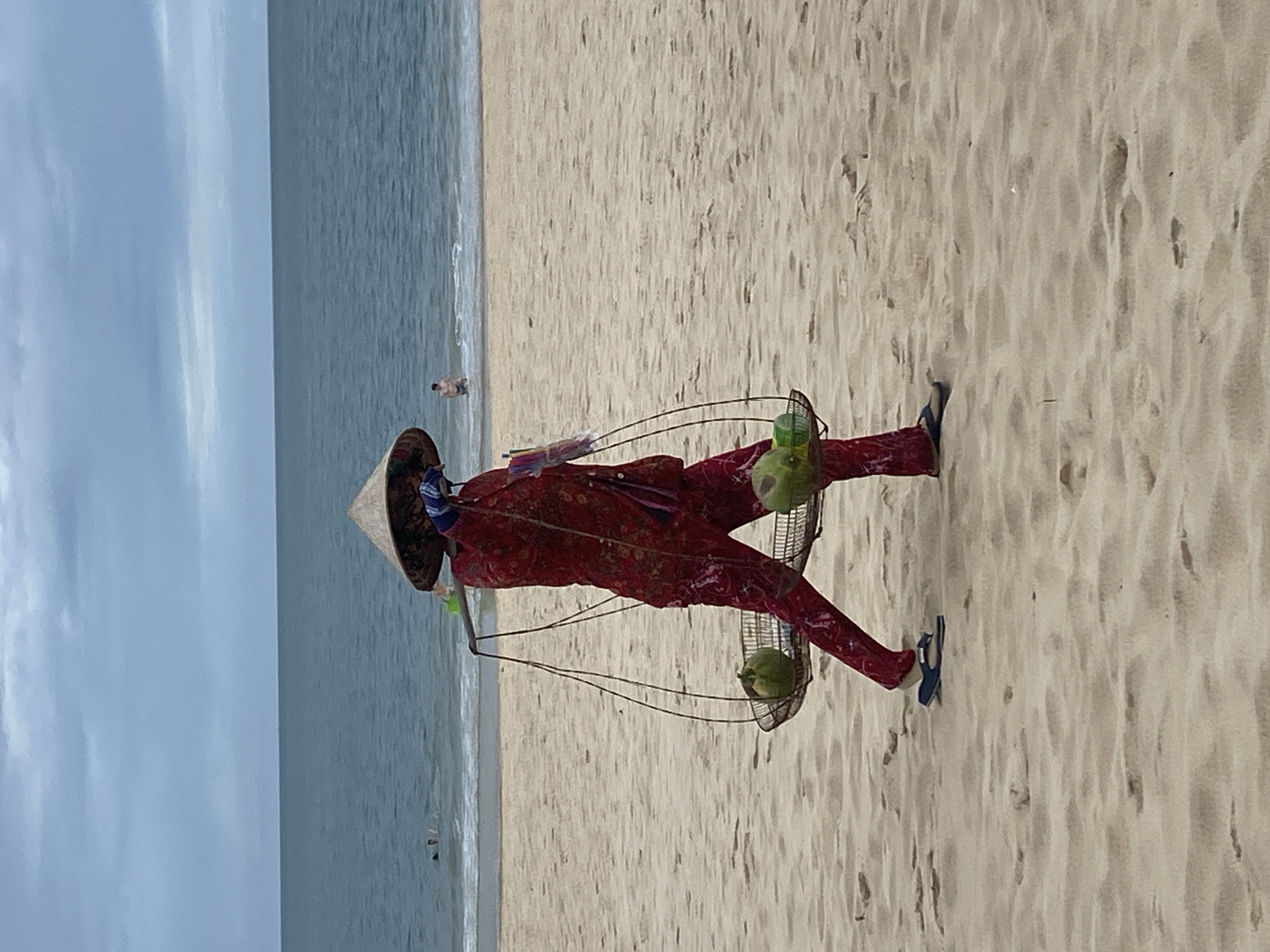 A person wearing traditional Vietnamese clothing and a conical hat stands on a sandy beach in Phu Thuy Ward, Vietnam. The person carries two wire baskets hanging from a shoulder pole, each containing a green coconut. The background includes the ocean and a cloudy sky. A few people are in the water.