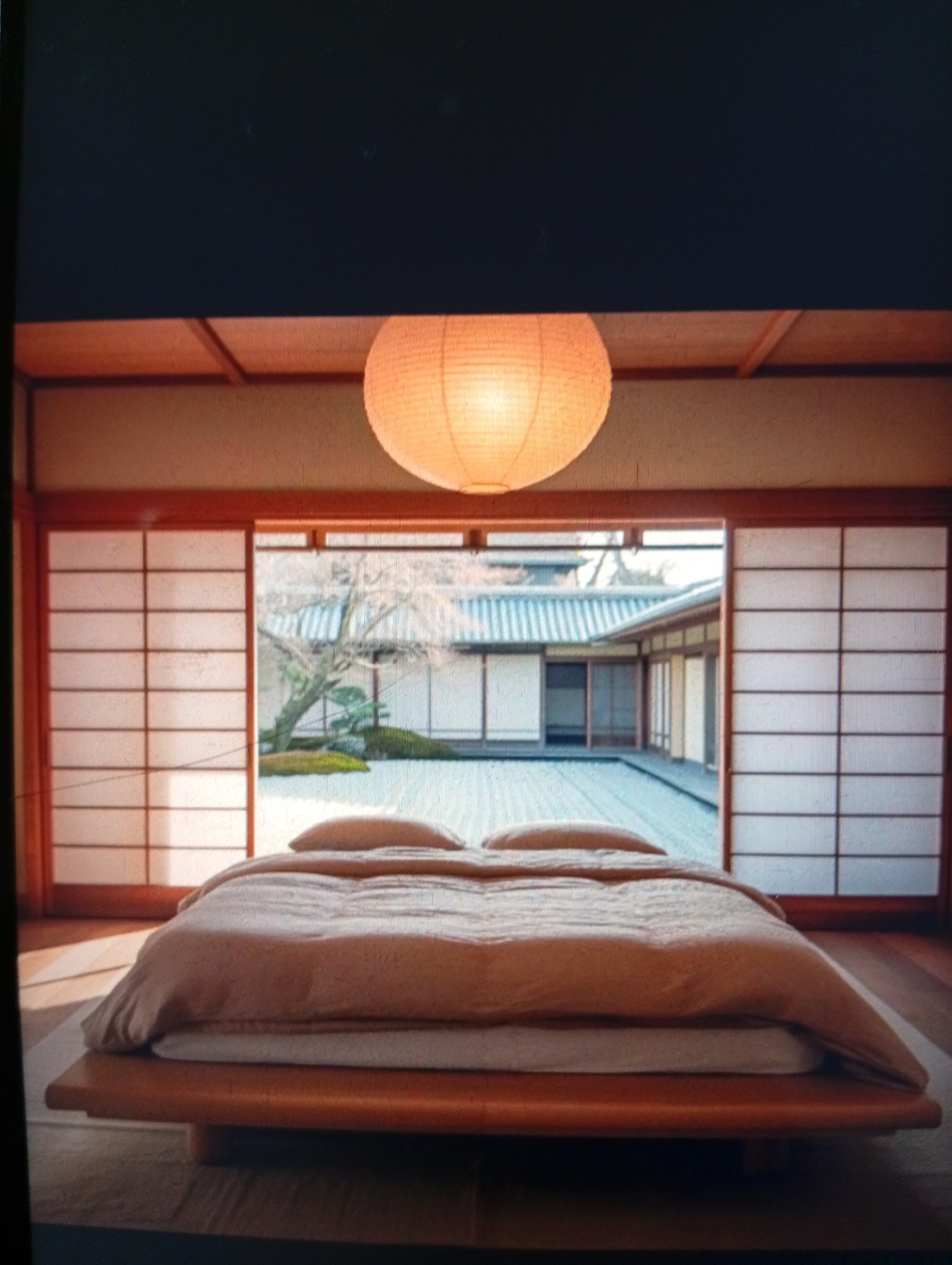 The image depicts a bedroom with a Japanese-style design. The bed, a low platform with a light peach-colored comforter, is positioned in the foreground. Above the bed hangs a round, orange paper lantern. Behind the bed, shoji screens flank a central opening that reveals a courtyard with a tree and building in the background. The building in the courtyard has a gray tiled roof. There are no people visible in the image. The location, Dutsen Alhaji, Nigeria, is not evident in the image.