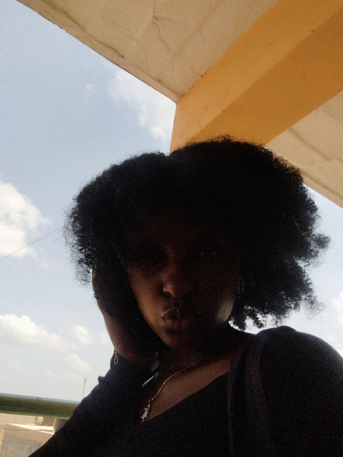 A close-up portrait, reportedly taken in Witeithie ward, Kenya, features a female individual. Her face and voluminous dark, curly hair are underexposed, appearing in silhouette against a bright, partly cloudy sky. She wears a dark-colored top and a silver-toned chain necklace with a pendant. Her right hand is positioned with fingers touching the area near her right ear. Her lips are pursed. Above and to the upper right of the subject, a light brown architectural ceiling or overhang and a structural beam are visible. A dark green railing is partially present in the lower-left background, with faint, indistinct structures in the distant background.