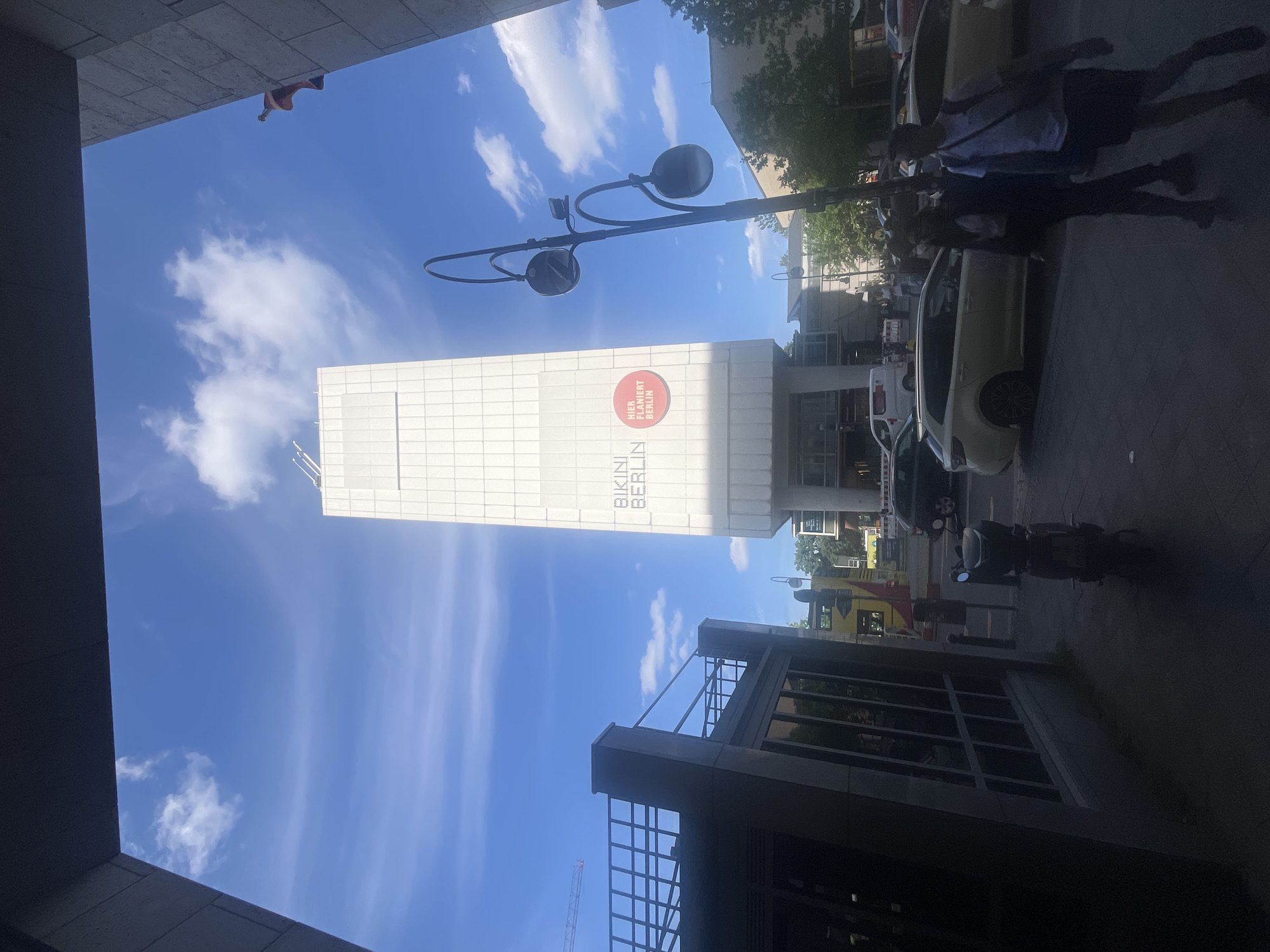 This is an outdoor, slightly upward-angled, full shot of a bright, sunny day in Berlin, Germany. The scene features a modern building with a white, grid-patterned facade that has "BIKINI BERLIN" written vertically on it. Above the text, a circular red logo with white text reads "HER FLANIERT BERLIN". A lamppost with two large black lights extends upwards into the blue sky dotted with wispy white clouds.
In the foreground on the right, a dark shadow is cast by a group of people, whose silhouettes are visible walking on the sidewalk. Their shadows are long, suggesting it's either morning or late afternoon. Next to them, a light-colored car is parked, and a dark motorcycle is also visible.
To the left and right of the frame, parts of buildings create a canyon-like effect, framing the sky and the central building. The overall impression is urban, with a focus on architecture and the interplay of light and shadow. The sky is clear and bright blue, indicating good weather conditions.