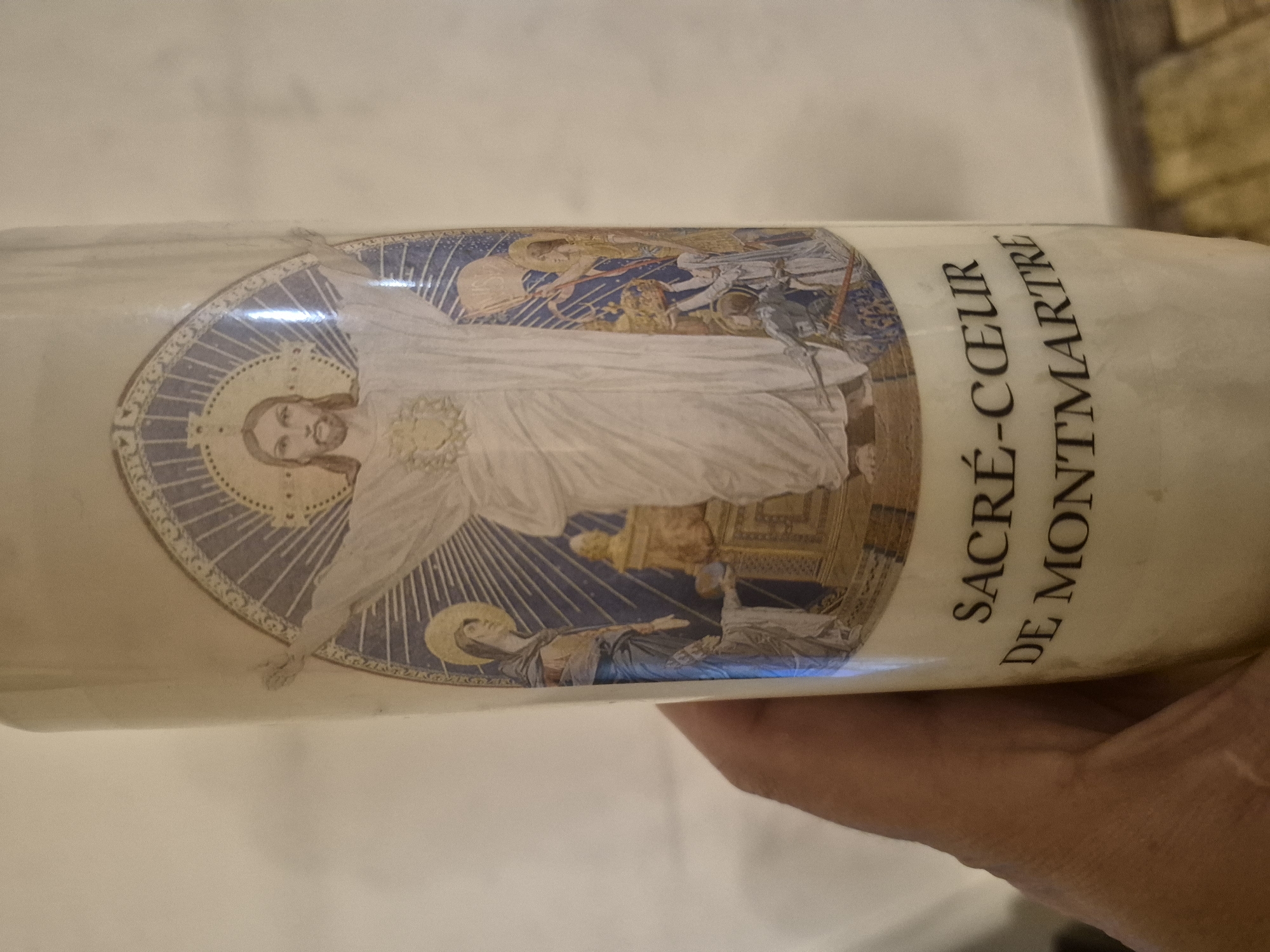 The image displays a large, cylindrical candle held by a human hand, presented in a slightly rotated view. The candle is adorned with a religious illustration featuring a central figure of Jesus Christ. Jesus is depicted with outstretched arms, wearing a white robe, and has a halo around his head from which rays of light emanate within a blue, star-filled circular motif. His Sacred Heart is visible, also emitting light.
Within the same circular frame, smaller religious figures are visible. Above Jesus's left shoulder (viewer's right), several figures, possibly saints or angels, are depicted in a heavenly scene. Below Jesus's right arm (viewer's left), a figure with a halo, likely Mary, is shown looking upwards, standing on a crescent moon. Another figure resembling a knight is also partially visible. The artwork style resembles classical religious painting or mosaic.
To the right of the illustration, prominently displayed in black text, are the words: "SACRÉ-CŒUR DE MONTMARTRE". This text strongly suggests the candle or its artwork is commemorative of, or originates from, the Basilica of the Sacré-Cœur in Montmartre, Paris, France. The scene beyond the candle appears to be an indoor setting with subtle background textures. The overall context is a religious devotional item, captured in Yerevan, Armenia.