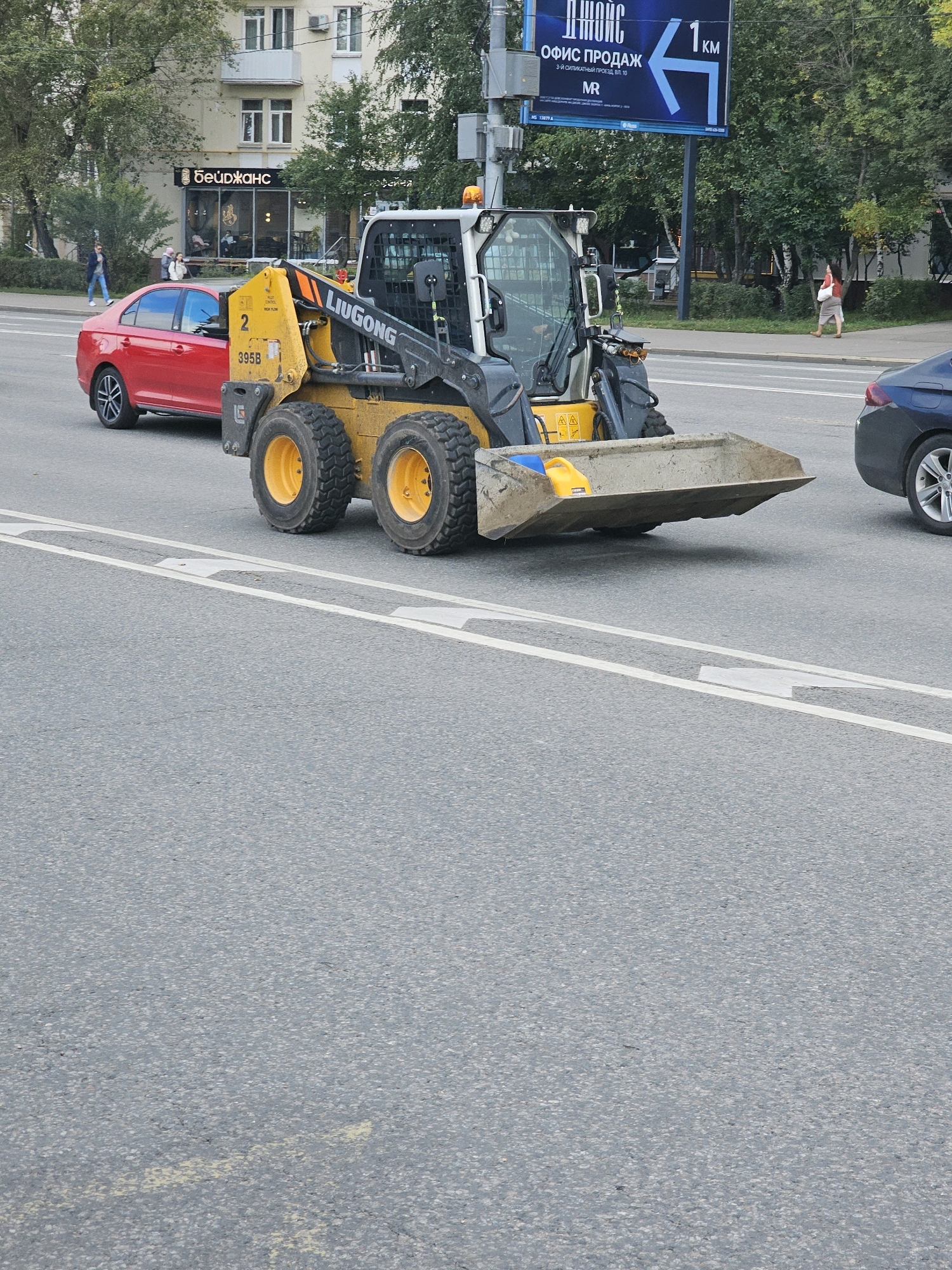 The image shows a street scene in Moscow, Russia. A yellow LIUGONG skid steer loader is parked on the road, facing right. The loader has the number '2' and '395B' visible on its side. A red sedan is parked to the left of the loader. In the background, there is a building with signs, including one with Cyrillic text that appears to be a business name. A pedestrian is walking on the sidewalk in the distance. A blue car is visible on the right side of the image. The road has white lane markings and there are trees and a traffic light pole in the background.