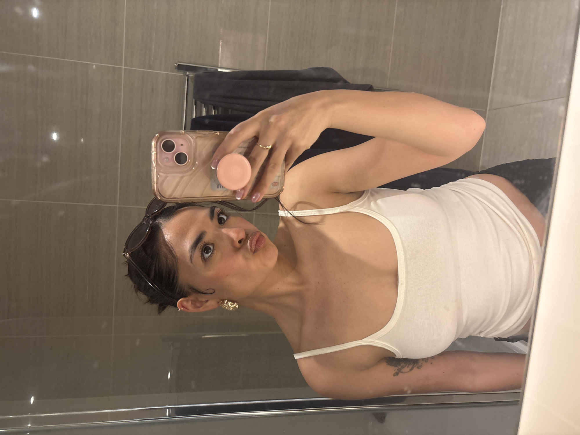 A woman is taking a selfie in a bathroom. She is holding a pink iPhone with a matching pink PopSocket. The woman is wearing a white tank top, gold earrings, and a gold ring. She has sunglasses pushed up on her head. The bathroom features gray tiled walls and a towel warmer with a dark-colored towel on it. The location is not definitively identifiable as being in London, United Kingdom.