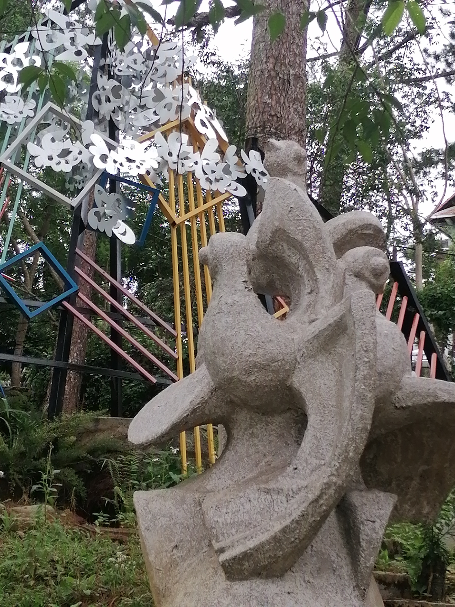 This is a medium shot, eye-level, outdoor photograph of a large, abstract stone sculpture of birds in flight. The sculpture is grey and textured, with flowing, organic lines. It is positioned in the foreground, slightly to the right of center. Behind the sculpture, a tall, rough-barked tree trunk is visible on the left. Further back, a series of colorful geometric frames, made of metal and painted in white, yellow, blue, and pink, are arranged in a cascading pattern, adorned with white paper cutouts that resemble butterflies or stylized flowers. These frames are supported by black metal posts. In the background, dense green foliage and more trees create a lush, natural setting. The ground in the foreground and midground is covered with a variety of green plants, including ferns and leafy bushes. The sky is overcast. The overall impression is one of art and nature integrated within an outdoor environment. This scene appears to be located in Da Lat, Vietnam, likely at a garden or art installation.