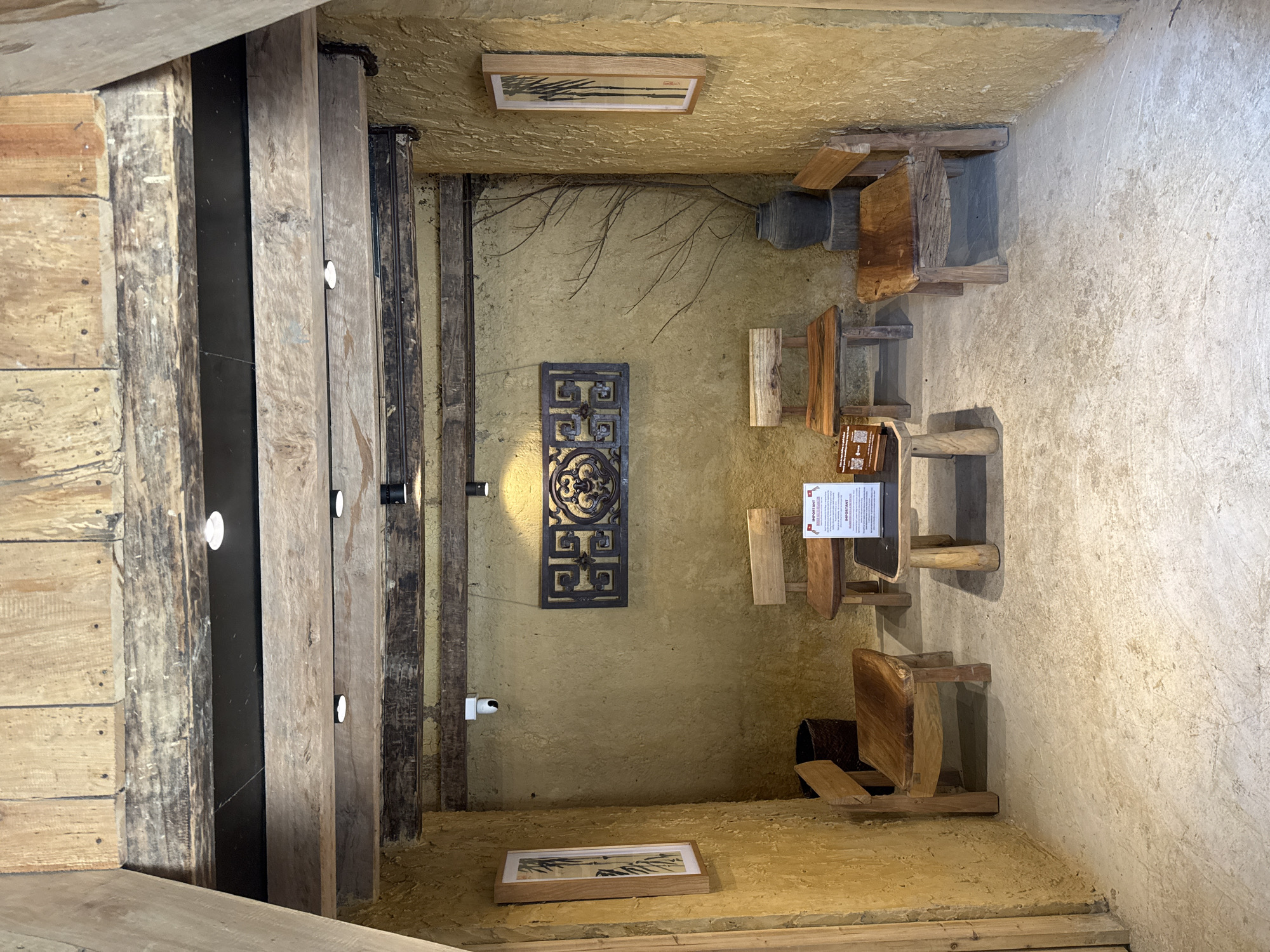 The image shows the interior of a building in Lũng Cú Commune, Vietnam. The walls are textured and yellowish-brown. There are wooden beams and planks along the left side, with embedded lights. A decorative metal panel hangs on the wall. Several small, rustic wooden stools and chairs are arranged in the room. Framed artworks are positioned high and low on the walls. A notice board stands on one of the stools.