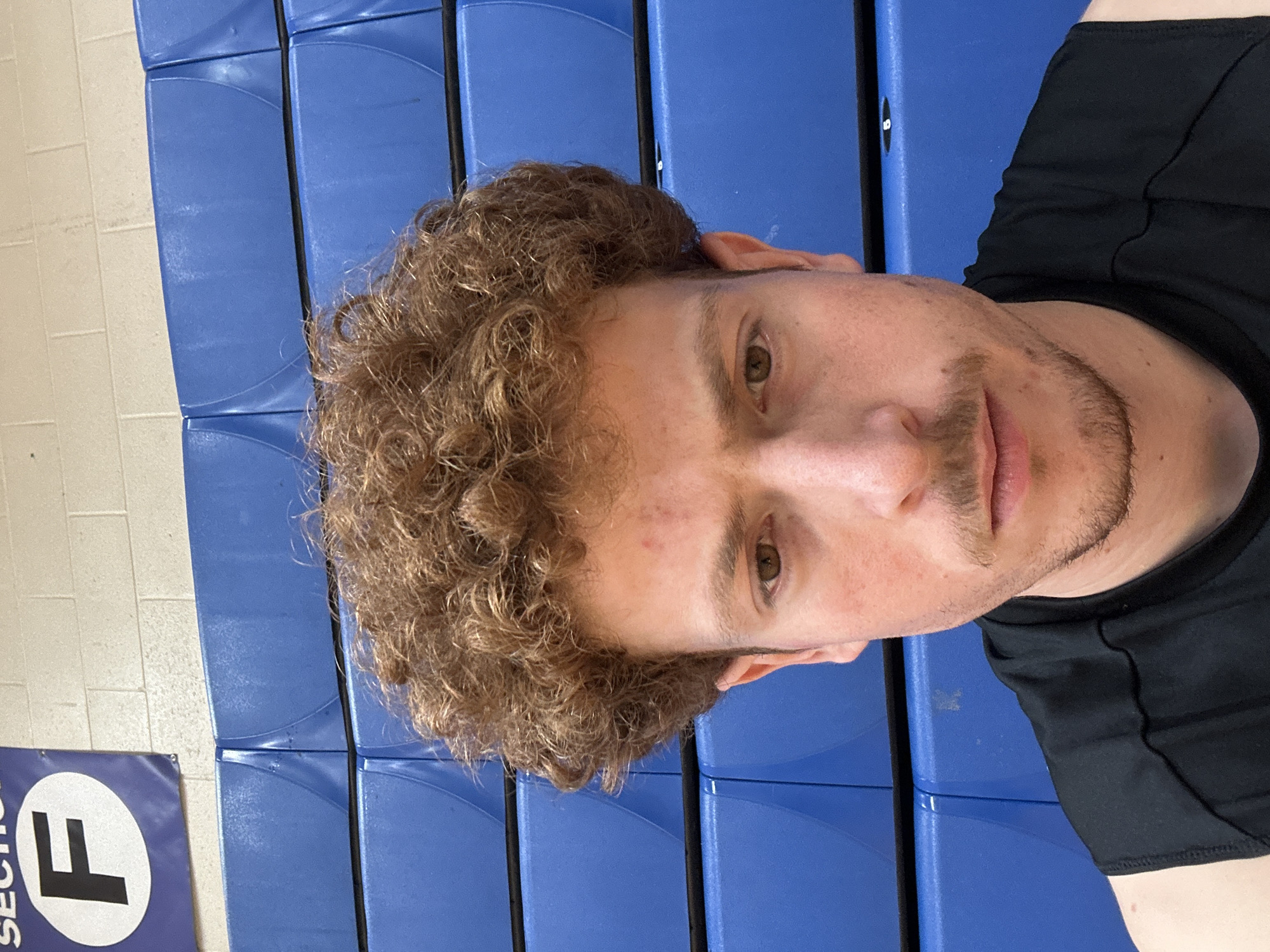 A close-up portrait features a light-skinned male with curly reddish-brown hair, brown eyes, and a light stubble on his upper lip and chin. He wears a black sleeveless athletic top and maintains a neutral, direct gaze. The background consists of multiple rows of cobalt blue plastic stadium seating, characteristic of an indoor sports venue or gymnasium. Above the seating, a light-colored brick or cinder block wall is visible. In the upper left corner, a blue banner with a white circle containing a black letter "F" and the partially visible word "SECTION" denotes a designated area. The environment is consistent with large indoor sports or entertainment facilities found in urban settings like New York, United States of America, though a precise location within the city is not discernible.