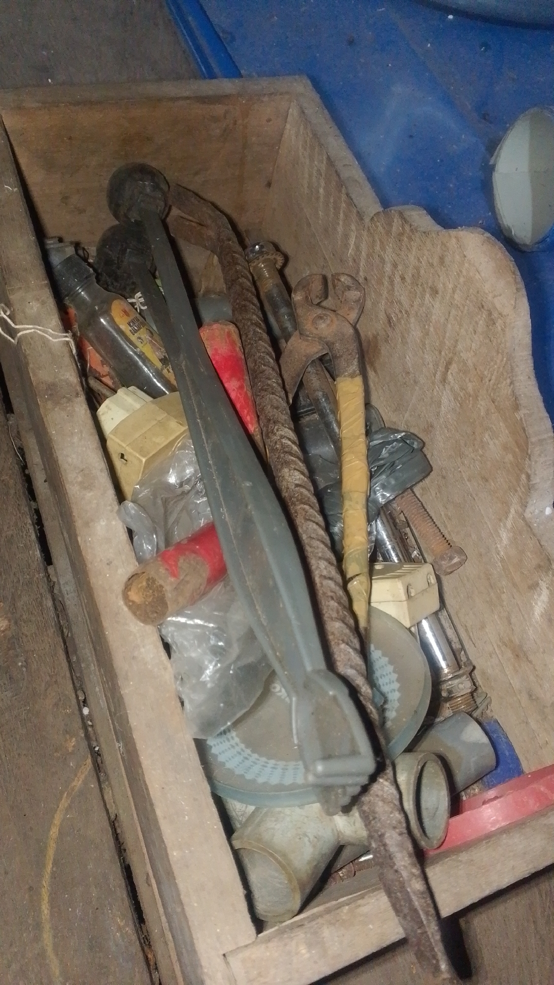 The image depicts a wooden toolbox filled with various metal tools. The tools appear old and some are rusty. Among the visible items are pliers, a hammerhead, a metal rod with a twisted pattern, a red-handled object, and what looks like a grinding wheel. Some of the tools are wrapped in plastic or tape. The toolbox itself is made of rough, unfinished wood and is placed on a wooden surface. The background is out of focus but shows a blue surface, possibly part of a vehicle or structure. The scene is dimly lit, suggesting an indoor or shaded outdoor environment. There are no people visible in the image. This image does not contain enough information to determine its location in Olak-Olak Kubu, Indonesia or any specific context.