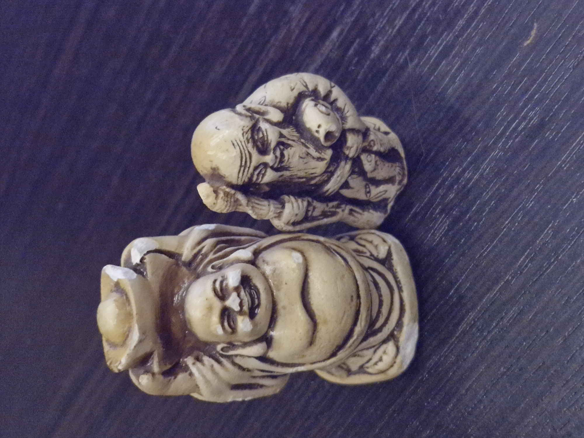Two small, decorative figurines are positioned side-by-side on a dark, textured surface. The figurine on the left depicts a laughing Buddha, holding a golden ingot above its head. It has a large, round belly, a cheerful expression, and is dressed in flowing robes. The figurine on the right is a representation of a wise old man, possibly a Taoist immortal. He has a long, flowing beard, wrinkled features, and is holding a staff in his left hand. He appears to be carrying a small object or creature against his chest. The figurines are made of a light-colored material, possibly resin or a similar composite, with carved details that suggest traditional East Asian artistry. The background surface exhibits a wood-grain texture with diagonal lines. The location is the city of Ерино, Russia.