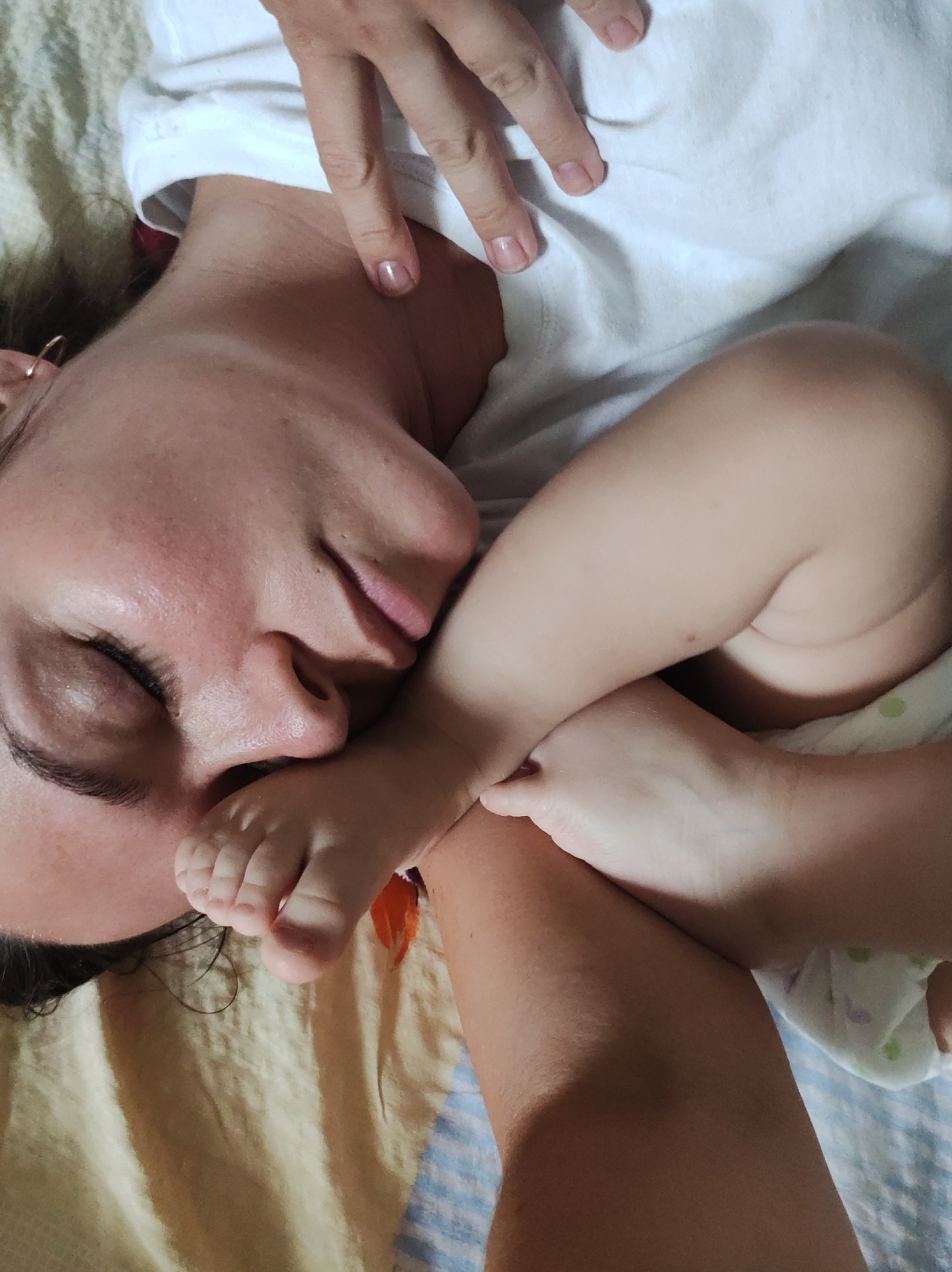 This close-up, top-down shot captures an intimate and tender moment between an adult and a baby, likely indoors in Фоки, Russia. The primary subject is an adult, presumably a woman, lying down with her eyes gently closed, suggesting rest or sleep. She is wearing a simple white t-shirt, and her dark hair is visible around her head. A small hoop earring can be seen on her left ear, and her fingers, adorned with clear nail polish, are resting near her shoulder.
Nestled close to the adult's face is a baby's tiny foot, reinforcing the bond between the two. Another chubby leg and thigh of the baby are visible, along with a glimpse of a white diaper featuring faint, light-colored patterns. The adult and baby are lying on a soft surface, which appears to be a bed or couch, covered by a textured, light-colored blanket, possibly off-white or light yellow, and a blue and white striped sheet or blanket towards the bottom right. The natural, soft lighting implies it is daytime. The scene evokes feelings of peace, comfort, and deep affection. There is no visible text in the image.