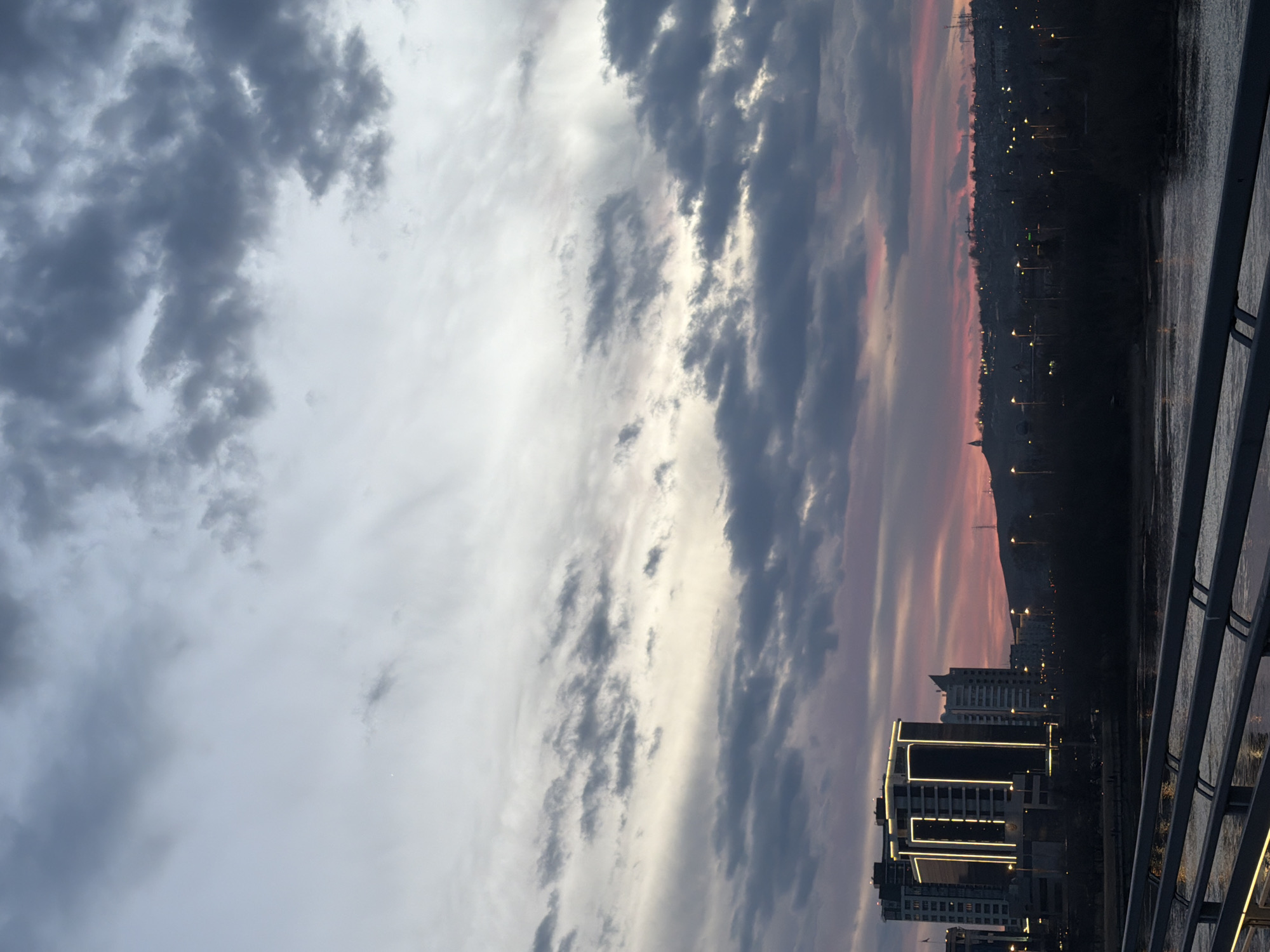 The image shows a cityscape at dusk in Krasnoyarsk, Russia. A modern building with distinctive illuminated lines is visible in the foreground. Behind the building are other residential towers, receding into the distance. The sky is filled with cloud cover, transitioning from grey on the left to pink and orange hues toward the right horizon. Lights are beginning to illuminate the city as twilight deepens. The view is likely captured from a bridge, indicated by the railing in the foreground, possibly across the Yenisei River. The image portrays a tranquil urban scene.