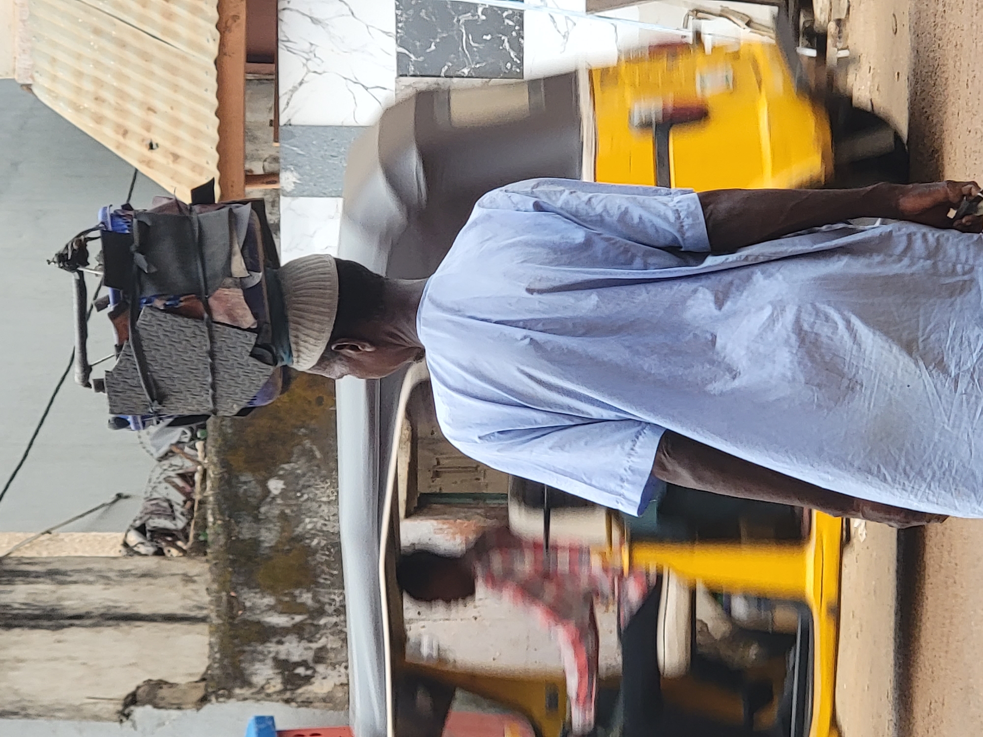 This vertical image, set in Maiburiji, Nigeria, captures an outdoor street scene under bright daylight.
The main subject is a dark-skinned man, positioned in the center, facing left in profile from the waist up. He wears a light blue, long-sleeved garment, likely a `kaftan` or `bubu`, and a light-colored skullcap (`kufi`). His right arm is visible, extended forward, and his hand is holding a small, dark object, possibly a phone or remote, with a ring on one finger.
Behind the man, to his right (from the viewer's perspective), is a yellow three-wheeled vehicle, characteristic of an auto-rickshaw or `keke napep`. The vehicle appears blurred, suggesting either motion or a shallow depth of field. A reflection of another person, wearing a red and white plaid shirt, is visible in the vehicle's window or surface.
The background features various architectural and environmental details. To the man's left, a pile of assorted items, possibly bags or textiles, rests on a structure that might be part of a stall or another vehicle. Above this, a corrugated metal roof, light brown in color, is visible. The wall behind the yellow vehicle is covered with white and grey checkered tiles, some exhibiting marble-like veining. A sturdy brown pole or beam is also present in the background. The ground appears to be a rough, unpaved surface.
The overall scene conveys a typical daily activity or interaction in an urban or semi-urban setting. The bright, even lighting indicates it is daytime. There is no visible text in the image.