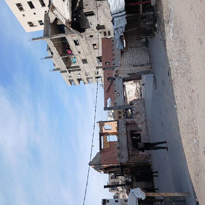 This image captures a scene of extensive urban destruction in Jabalia, Palestinian Territory, under a partly cloudy blue sky. The foreground consists of a dirt and gravel road leading into a wider, flat area of compacted earth scattered with debris. Three individuals are visible: a person in a black hoodie stands centrally, facing away from the camera, observing the damaged environment. Two additional figures are partially obscured in the left background, near a severely damaged building.
The midground is dominated by the skeletal remains of multiple structures. A prominent reddish-orange colored concrete building exhibits large missing sections of its facade, exposing interior spaces and structural elements, and bears numerous circular impact holes across its remaining walls. To its right, another building features a partially collapsed roof and extensive wall damage. A partial stone-clad wall with an arched opening stands on the right, also showing damage.
In the background, a multi-story concrete building, appearing either under construction or heavily damaged, shows exposed rebar, missing wall sections, and articles of laundry hanging from balconies or exposed floors. Other multi-story buildings are partially visible further in the background. A black utility line with a single light fixture stretches horizontally across the middle ground. On the far right, a makeshift market stall structure covered by white and blue tarpaulins is present.