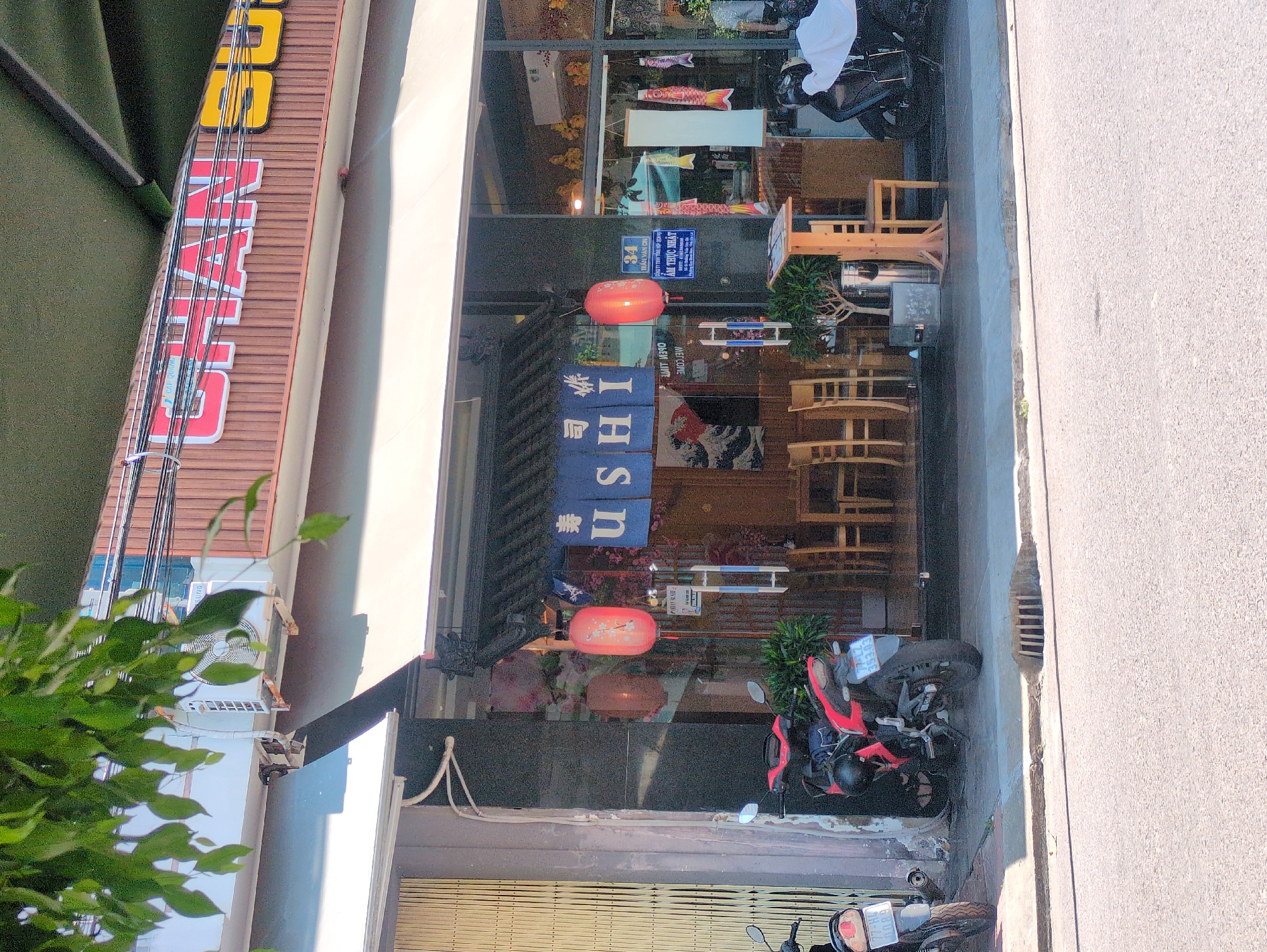 The image shows a vertical, eye-level, outdoor view of the front of a Japanese sushi restaurant in Quy Nhon Ward, Vietnam. The restaurant has a large, vertical, multi-colored sign, partially obscured by a green tree in the lower left. The sign reads “SUNG” at the top, followed by a series of Japanese characters, and “USHI” in Roman letters. The restaurant's entrance is decorated with Japanese lanterns and banners. Inside, wooden tables and chairs are visible. Two motorcycles are parked in front of the restaurant on the sidewalk. A sign that reads, “34” at the top with Vietnamese text below is affixed to the glass window. A concrete sidewalk and road are visible to the right.