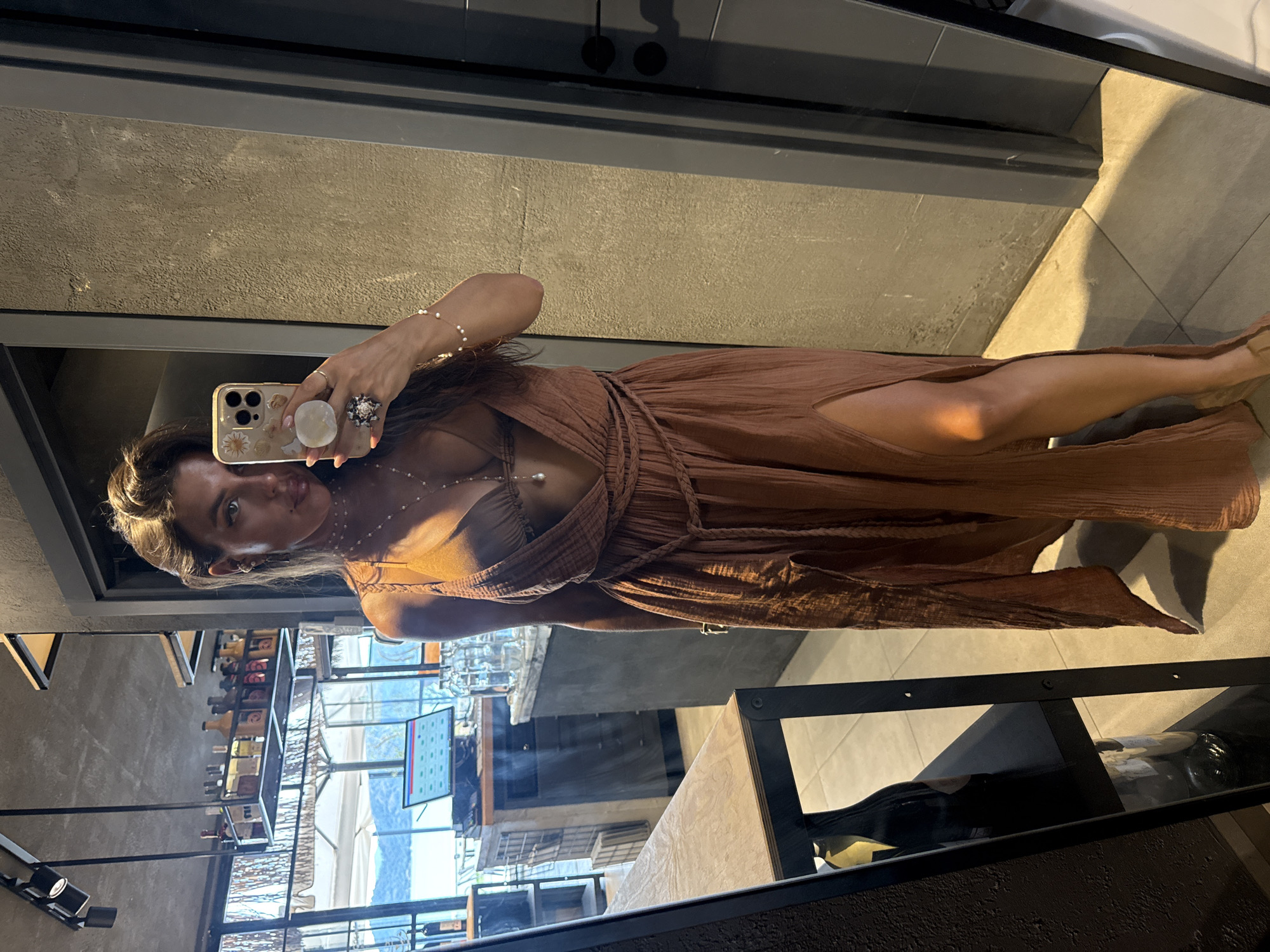 A woman stands in front of a mirror, taking a selfie. She is wearing a long, brown, flowy dress with a deep V-neck and a slit up the side, revealing her leg. She has on a bikini top underneath the dress, and several delicate necklaces. She is also wearing rings and a bracelet. Her hair is dark and pulled back. The background appears to be an indoor establishment, possibly a restaurant or bar, with shelves stocked with bottles. Through a large window in the background, a body of water and hills can be seen, suggesting a coastal location. The overall impression is that the photo was taken in Fethiye, Turkey, likely during a vacation.