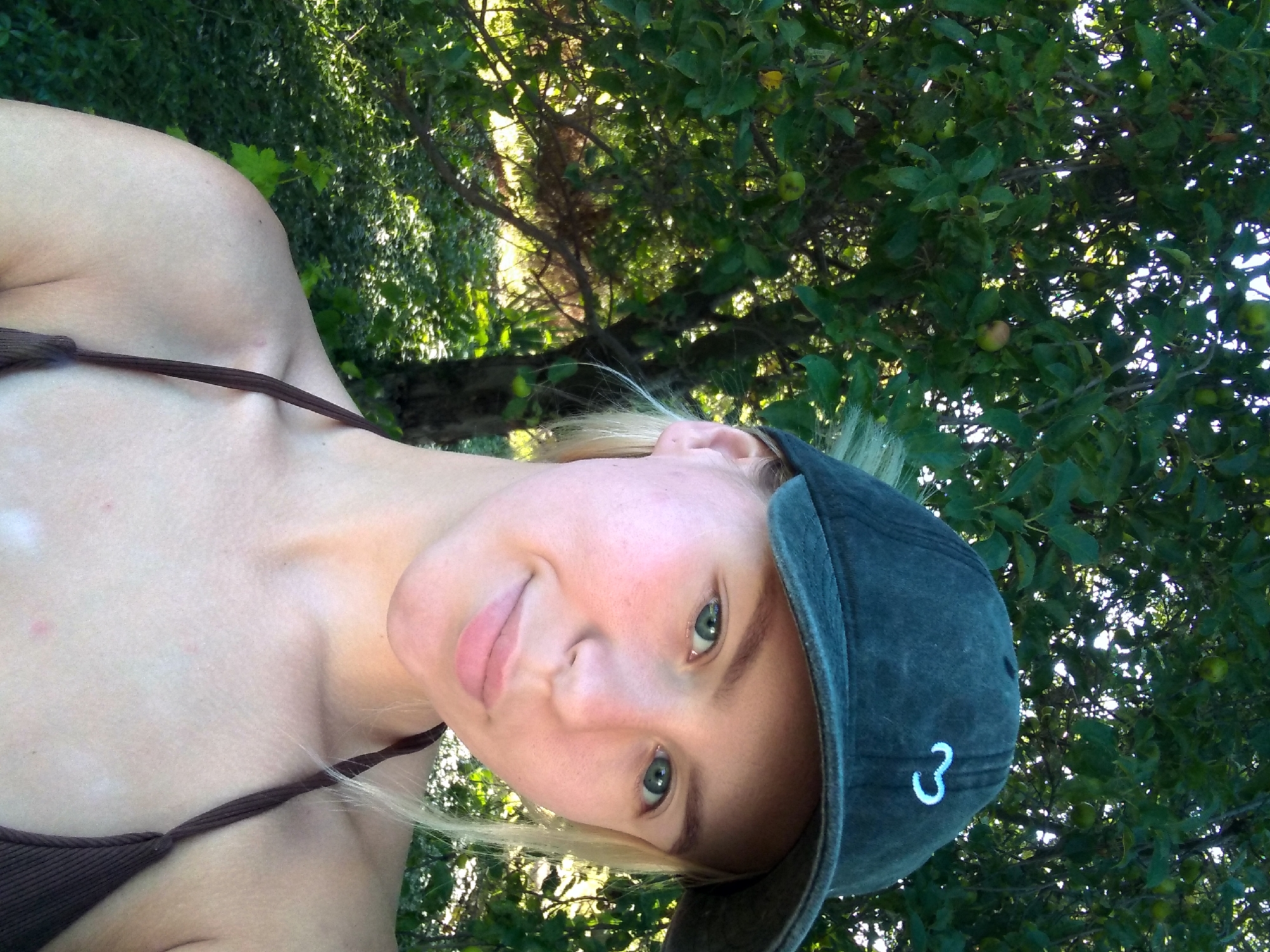In Radisne, Ukraine, a young woman with blonde hair pulled back from her face and wearing a dark baseball cap and a brown bikini top poses for a selfie. She is positioned in front of a tree with green leaves and some small, green fruits, suggesting an orchard or a garden setting. The sunlight filters through the branches, creating a dappled effect on the background foliage. The woman's face is turned slightly towards the camera, and she has a subtle smile. Her skin appears fair, and her eyes are blue-green. The overall impression is a candid outdoor portrait, likely taken during a warm, sunny day.