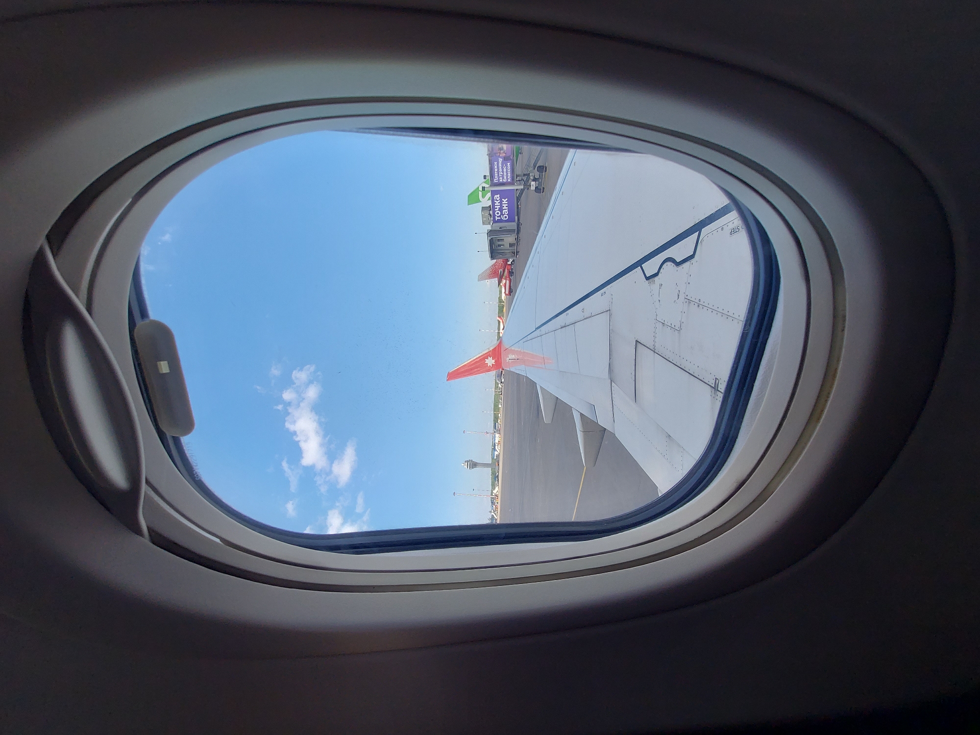 View from an airplane window. A white airplane wing with visible rivets is visible. The red tail of another airplane with a white star is visible. In the background, an airport control tower and several airport service vehicles are visible. Advertising for the "Tochka Bank" is visible on one of the service vehicles. The sky is blue with a few clouds. The window frame is light gray. The plane is on the ground at Pulkovo Airport in Saint Petersburg, Russia.