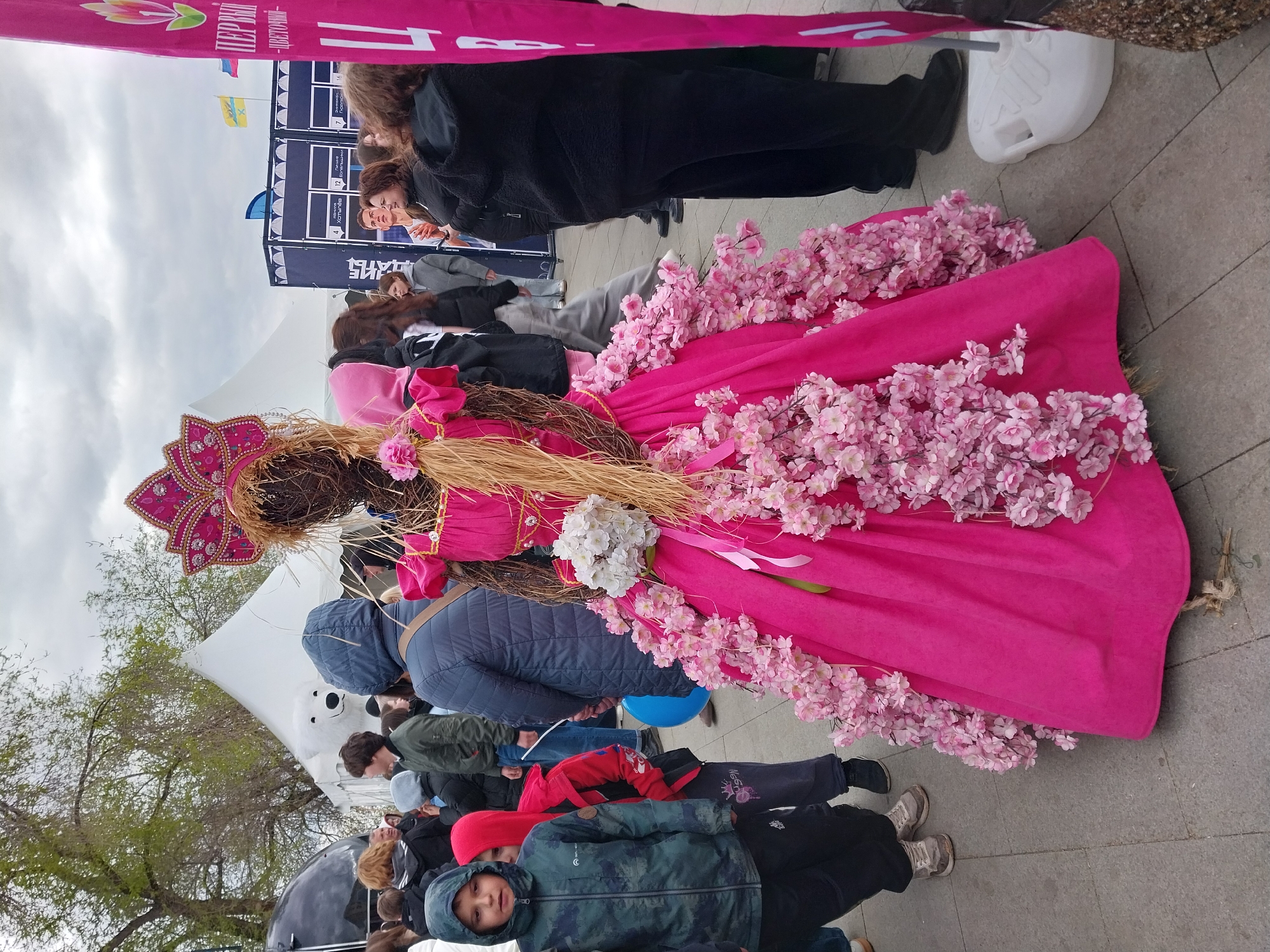 A festival event is captured in a medium outdoor shot, likely in Orenburg, Russia. The focal point is a large, ornamented straw doll. The doll is dressed in a vibrant pink garment with pink flowers and is adorned with a decorative headdress.
The doll sits on the ground among a group of people, including children, with individuals standing and sitting around it. Several buildings and tents, along with banners displaying Russian text, are visible in the background. The ground beneath the doll is paved with grey stone, adding a formal and public character to the scene.