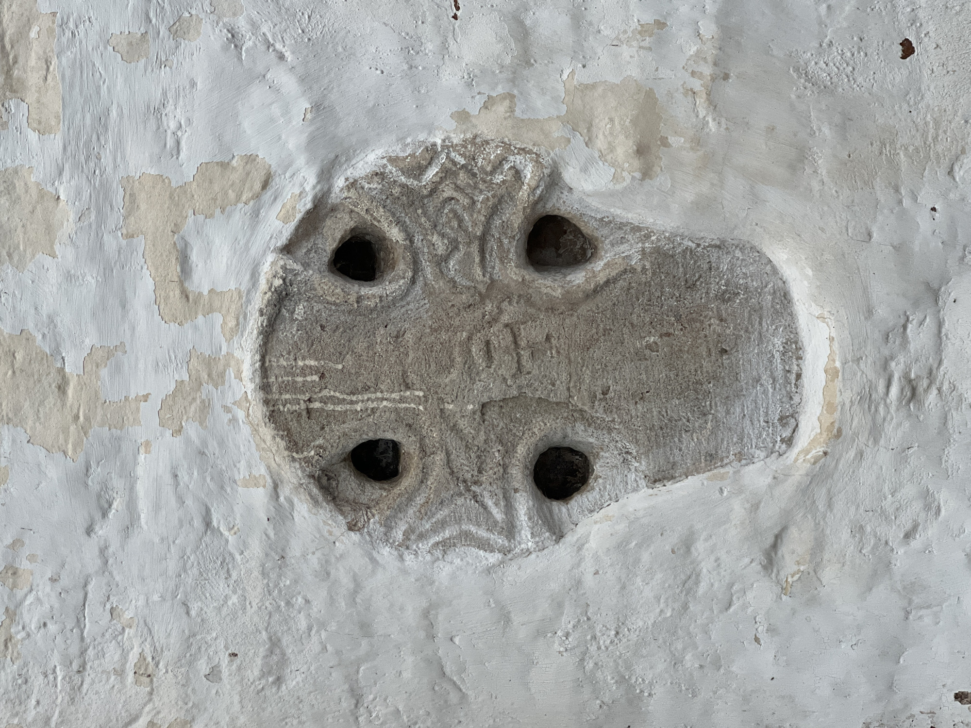 The image shows a close-up, weathered stone carving embedded in a white, crumbling plaster wall. The carving is shaped like a cross with four circular holes, one at the intersection of the arms and one at the end of each arm. The stone has intricate, worn carvings on its surface, depicting swirling patterns and what appear to be stylized animal or abstract motifs. The plaster wall surrounding the carving is heavily damaged, with large flakes of paint peeling away to reveal the underlying brickwork or rough plaster. The color palette is muted, consisting of shades of white, beige, and gray. The lighting is flat, suggesting an overcast day or indoor setting, which highlights the texture and imperfections of the materials. There are no people or other objects visible in the frame. This stone carving is likely an architectural detail or a historical artifact.