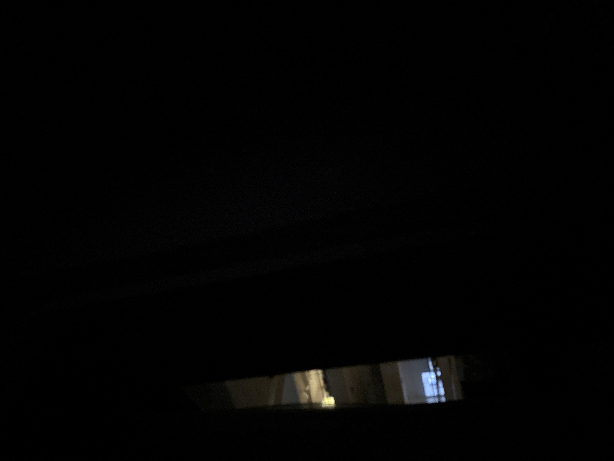 The image is a dark and unclear view through a small opening. The light from the opening reveals an interior space, potentially a hallway or room, with light reflecting off surfaces within. It's impossible to discern specific objects or people due to the poor visibility, but the background suggests a building interior. The information required to determine the location in the city of Budva, Montenegro is not available.