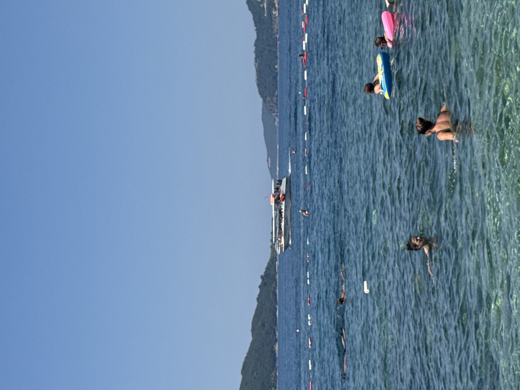 The media file depicts a sunny day at a beach in Kamenovo, Montenegro. The scene is split between a clear blue sky on the left and a vibrant blue sea on the right. The water is calm, with visible ripples and streaks of light reflecting off the surface.
Several people are enjoying the water. Some are swimming, while others are lounging on inflatable toys. In the foreground, a person is partially submerged, with only their head and shoulders visible above the water. Further out, a pink inflatable raft and a blue inflatable ring are being used by individuals. Closer to the shore, a boat with several people on deck is visible, suggesting a tour or a private vessel.
A line of buoys marks a boundary in the water, separating different areas of the sea. The shoreline is lined with lush green trees and vegetation, indicating a mountainous landscape behind the beach. The overall atmosphere appears relaxed and recreational, consistent with a typical beach day. The lighting suggests it is daytime, likely midday or early afternoon, with bright sunlight illuminating the scene. There is no visible text in the media file.