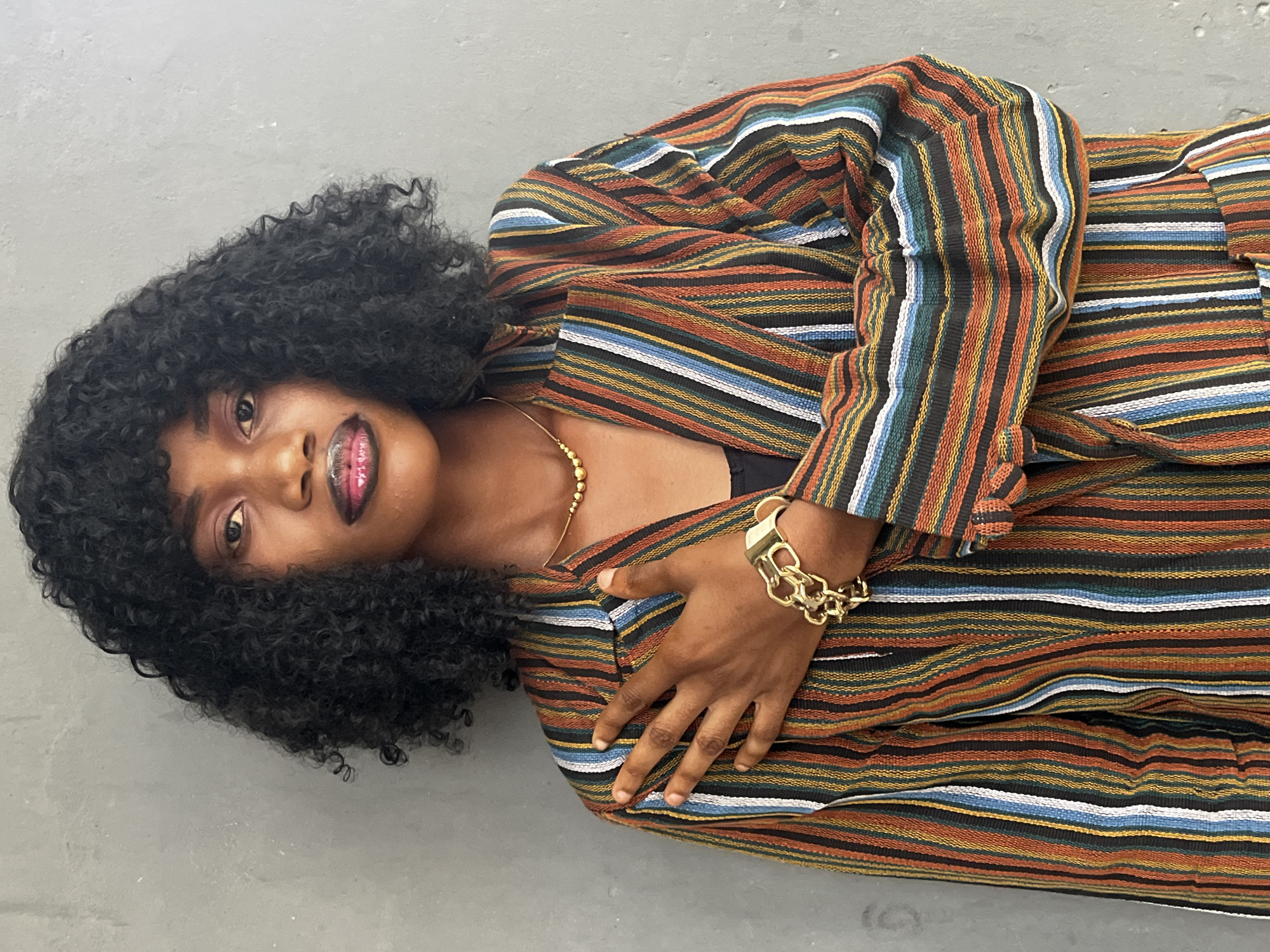 A Black woman with curly black hair is lying on a gray surface. She is wearing a striped blazer with orange, blue, white, and brown stripes, a gold necklace, and a large gold bracelet. She has dark lipstick and is looking towards the camera. The location is not specified.