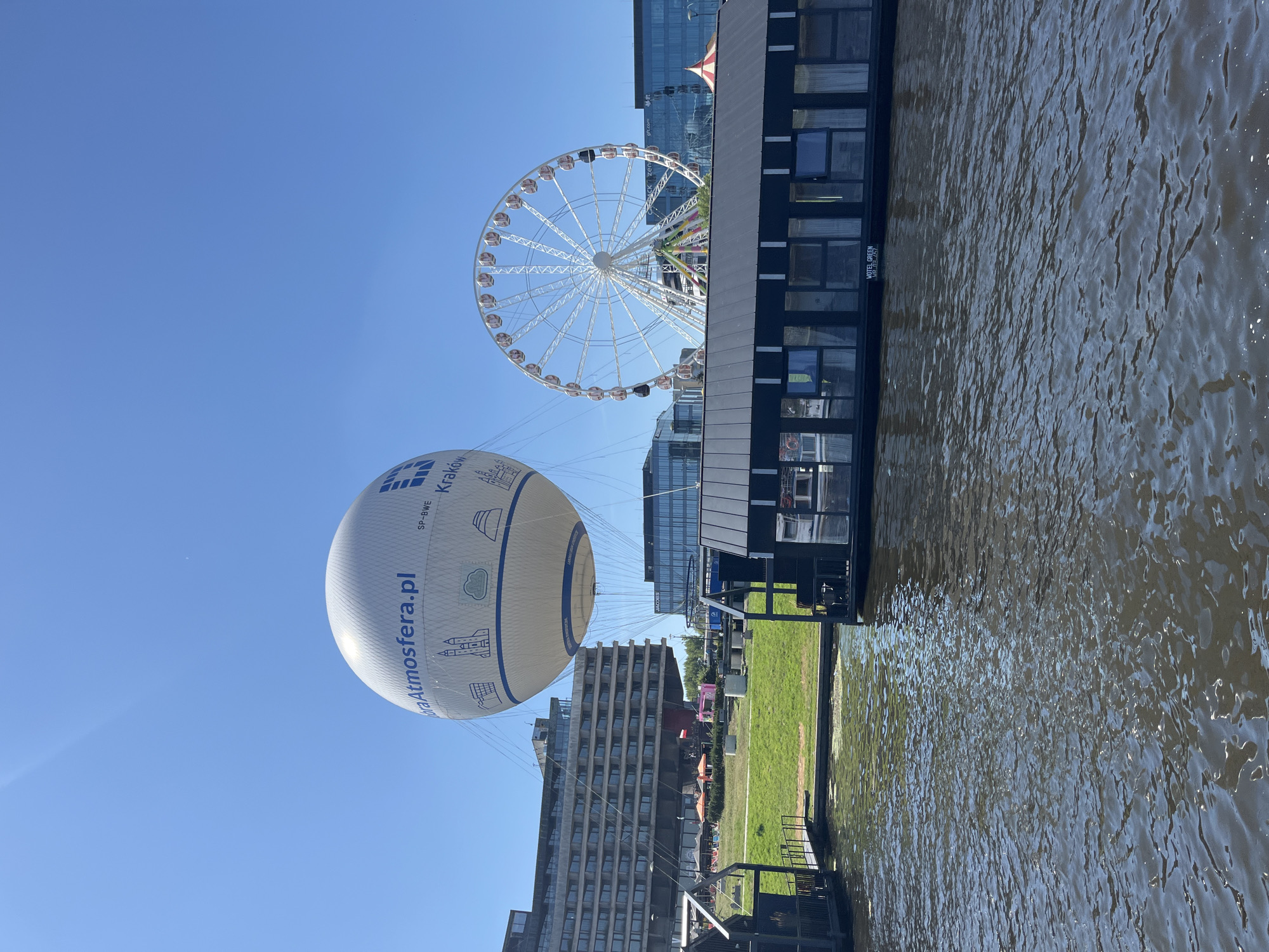 An aerial view shows a large white hot air balloon tethered in the sky, with the website "Atmosfera.pl" and "Kraków" visible on its side. To the right of the balloon, a Ferris wheel stands tall, its cabins a mix of white and pink. In the foreground, a dark, modern building rests on the water, its rectangular windows reflecting the blue sky. The building appears to be a hotel, with "HOTEL GREEN" printed on its side. The water is a deep, dark color, rippling gently. Behind the hotel, a lush green lawn stretches towards a row of modern buildings. The sky is a clear, bright blue, indicating a sunny day. The scene is set in Krakow, Poland, likely near a waterfront area with recreational attractions. There are no people visible in the image, and no actions or interactions are occurring.