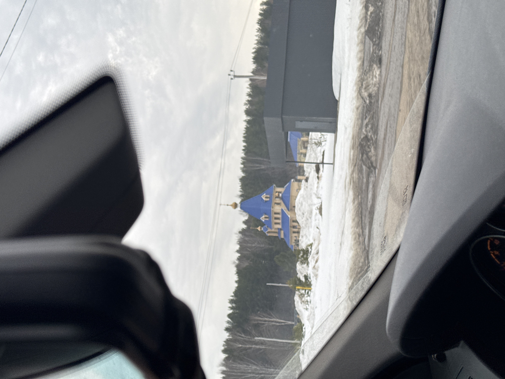 View from inside a vehicle, possibly a car, looking out the front windshield, with a rear view mirror partially visible on the left. The outside view shows a snow-covered landscape with a forested area in the background and a building with blue roofing. The location is likely in Chaikovsky, Russia, but the precise location is undetermined.