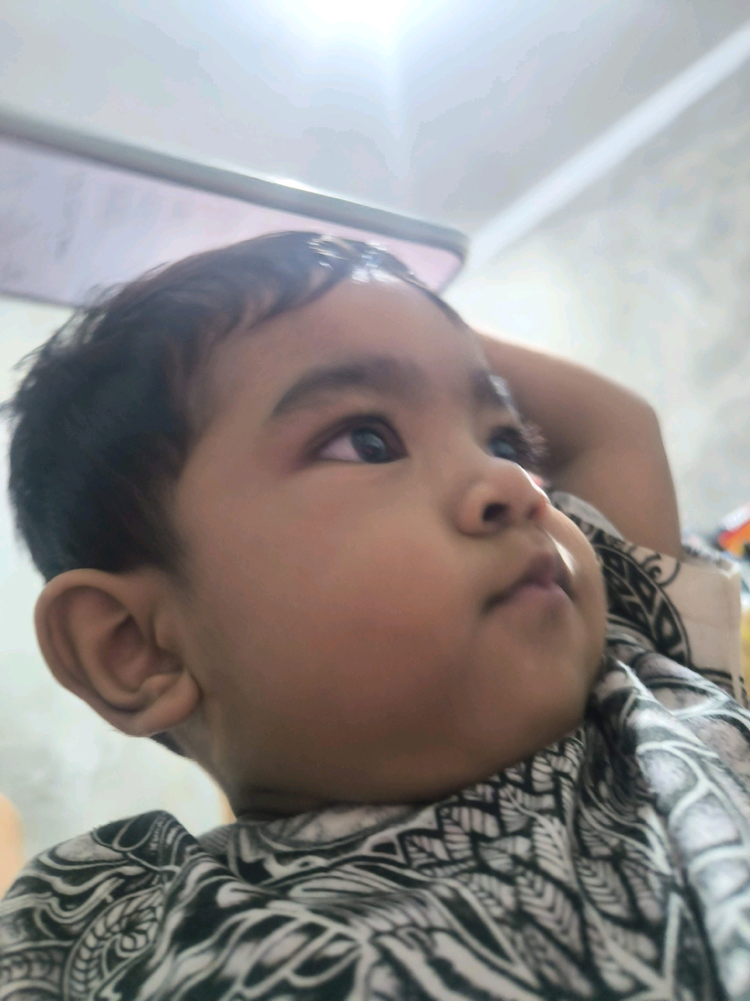 The image is a close-up, low-angle shot of a young child's face, positioned in Tabanan, Indonesia. The child, with dark hair and eyes, is looking upwards and to the right. Their right arm is raised and bent, with their hand resting behind their head. They are wearing a black and white patterned garment. The background is out of focus, showing a ceiling with a light fixture. The overall lighting suggests an indoor setting.