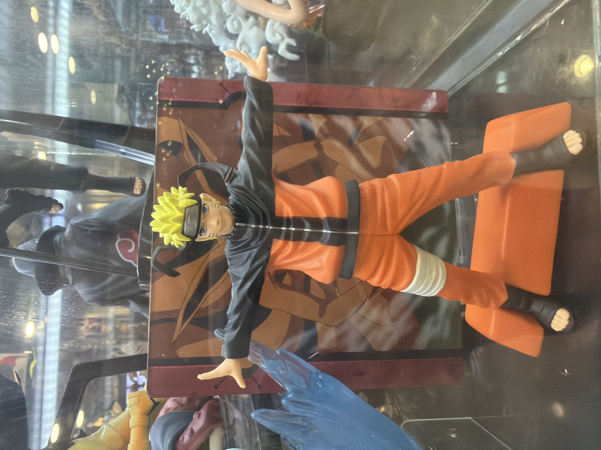 A Naruto Uzumaki action figure stands in a display case, dressed in his signature orange jumpsuit and black jacket. The figure has spiky blonde hair and blue eyes. Behind the figure is a large, orange and brown backdrop featuring the Uchiha clan symbol. To the left of the figure, a translucent blue water-like effect is visible. The figure is positioned on an orange platform. The setting appears to be a retail store or collector's shop, possibly in Davao City, Philippines, given the potential context implied by the prompt.