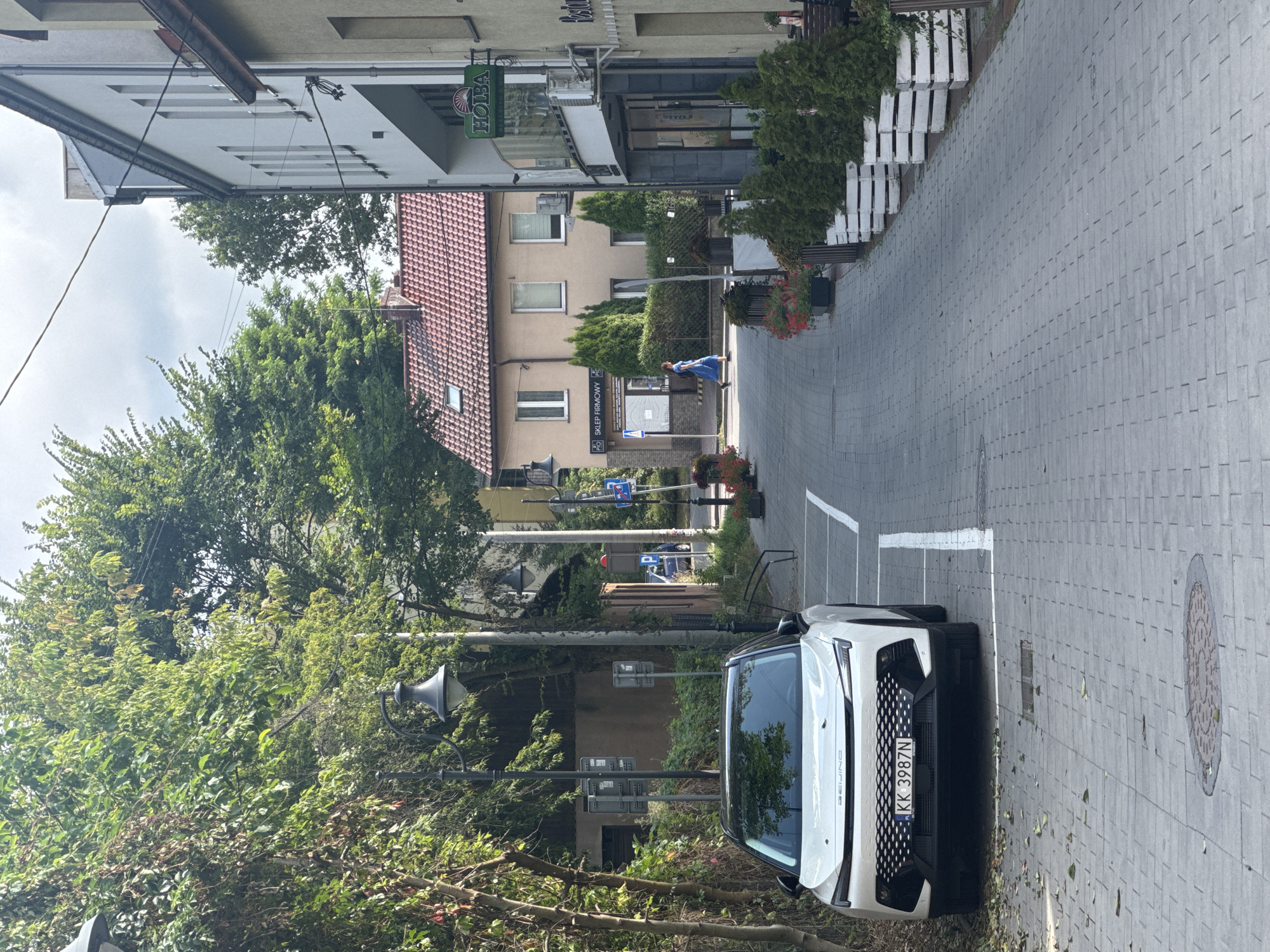 A white electric car, a BEIJING brand, is parked in a designated spot on a cobblestone street. The car's license plate reads "KK 3987N". In the background, a woman in a blue dress walks down the street. The street is lined with buildings, trees, and planters. One building has a sign that says "SKLEP FIRMOWY". Another building has a sign that says "HOLBA". The sky is overcast. The scene appears to be in a town or city, possibly in Eastern Europe based on the license plate and architecture.