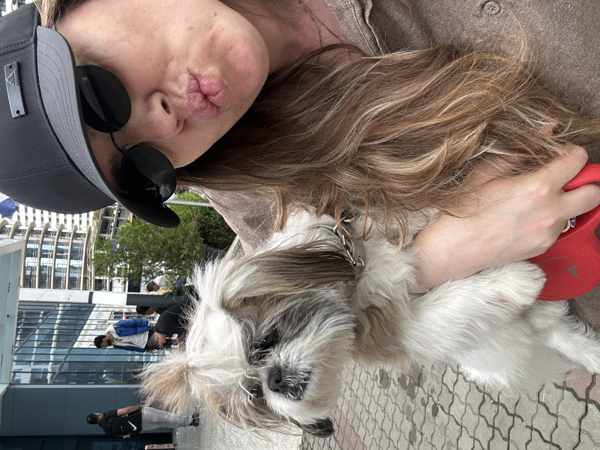 A medium shot shows a woman in a brown shirt, black baseball cap, and black sunglasses holding a Shih Tzu dog. The woman is puckering her lips for a selfie. The dog is fluffy and white with brown and gray markings, and is wearing a silver chain collar. The woman's right hand, which is holding the dog, is also holding a red dog leash. In the background, several people are standing outside a modern building with large glass windows. The location appears to be an urban area, possibly Batumi, Georgia, given the contemporary architecture. The ground is paved with gray interlocking bricks.