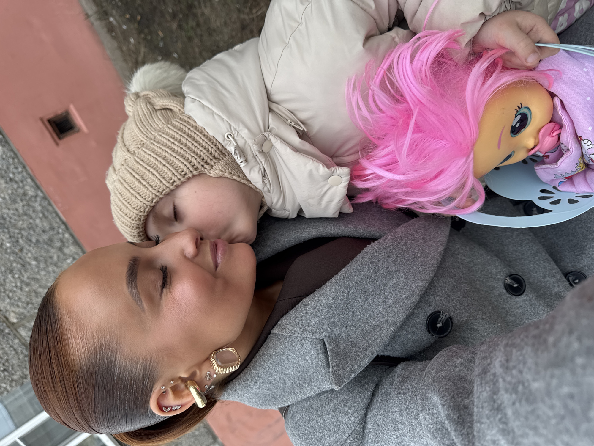 The image depicts an adult female and a young child engaged in an affectionate interaction within the city of Chaikovsky, Russia. The woman, positioned on the left, has brown hair pulled back and is wearing a grey overcoat over a dark brown garment. Her left ear displays multiple piercings, including a gold-toned ear cuff, several small clear studs, and a large gold-toned earring with an oval, clear center. Her eyes are closed as the child, wearing a beige knitted beanie with a white pom-pom and a light beige puffy winter jacket, presses their lips against her left cheek. The child's left hand is holding a light blue plastic doll carrier. Inside the carrier is a doll with bright pink hair, a yellow-skinned face, large blue eyes, and a pink pacifier in its mouth, dressed in a light purple garment. The background consists of a plain, faded pink building wall with a small, dark rectangular window visible in the upper right. Below the wall, a patch of light brown ground with sparse white specks is present.