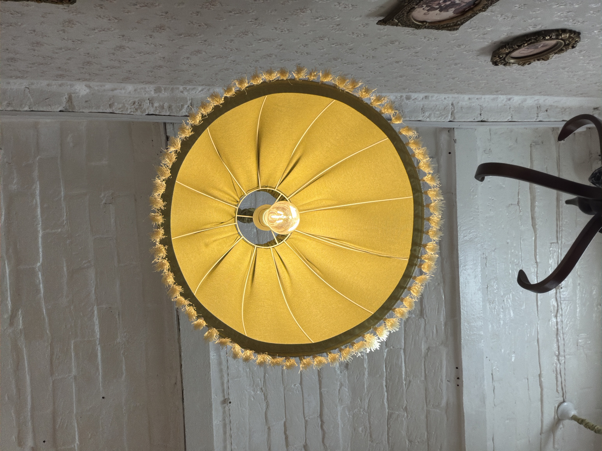A low-angle shot depicts a mustard-colored lamp hanging from a textured white ceiling. The lamp shade is circular with gathered fabric and a decorative fringe. A lightbulb illuminates the space beneath the shade. The surrounding walls are painted white and appear to be constructed of brick. Two framed pictures are visible on the ceiling towards the top of the frame. A dark-colored decorative element appears on the right side of the frame. The location is Sarapul, Russia.